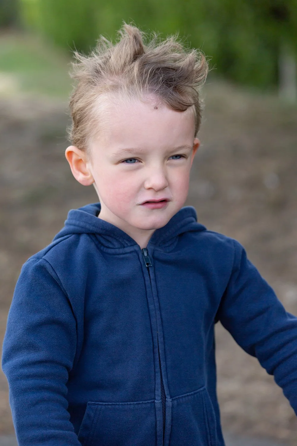 Young boy with tousled hair wearing a navy blue hoodie outdoors with blurred trees in the background.