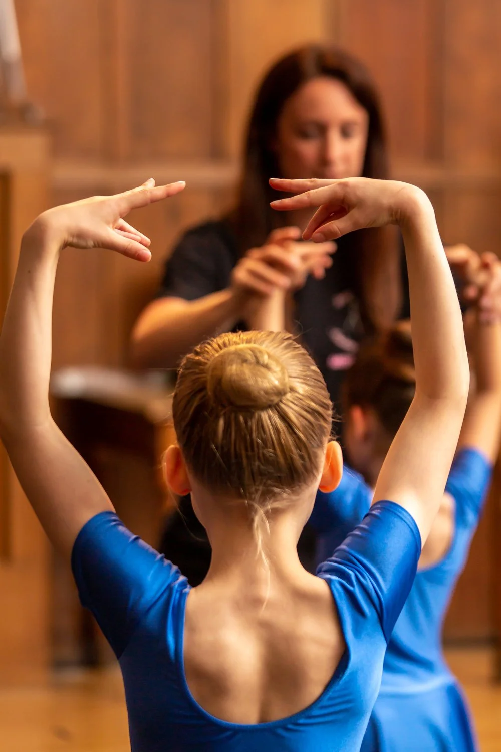 A young ballet dancer with her hair in a bun posing with arms raised, facing an instructor or coach in a dance studio.