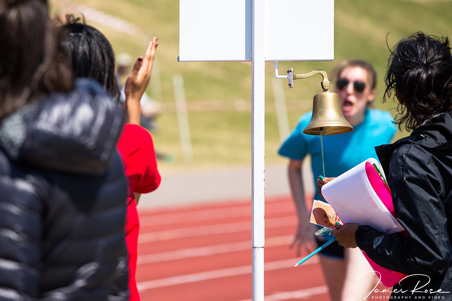 An award bell at a race event with people in the background, including a woman with black hair holding a notebook and a pen, and another individual in a blue shirt speaking or shouting, on a track field during daytime.