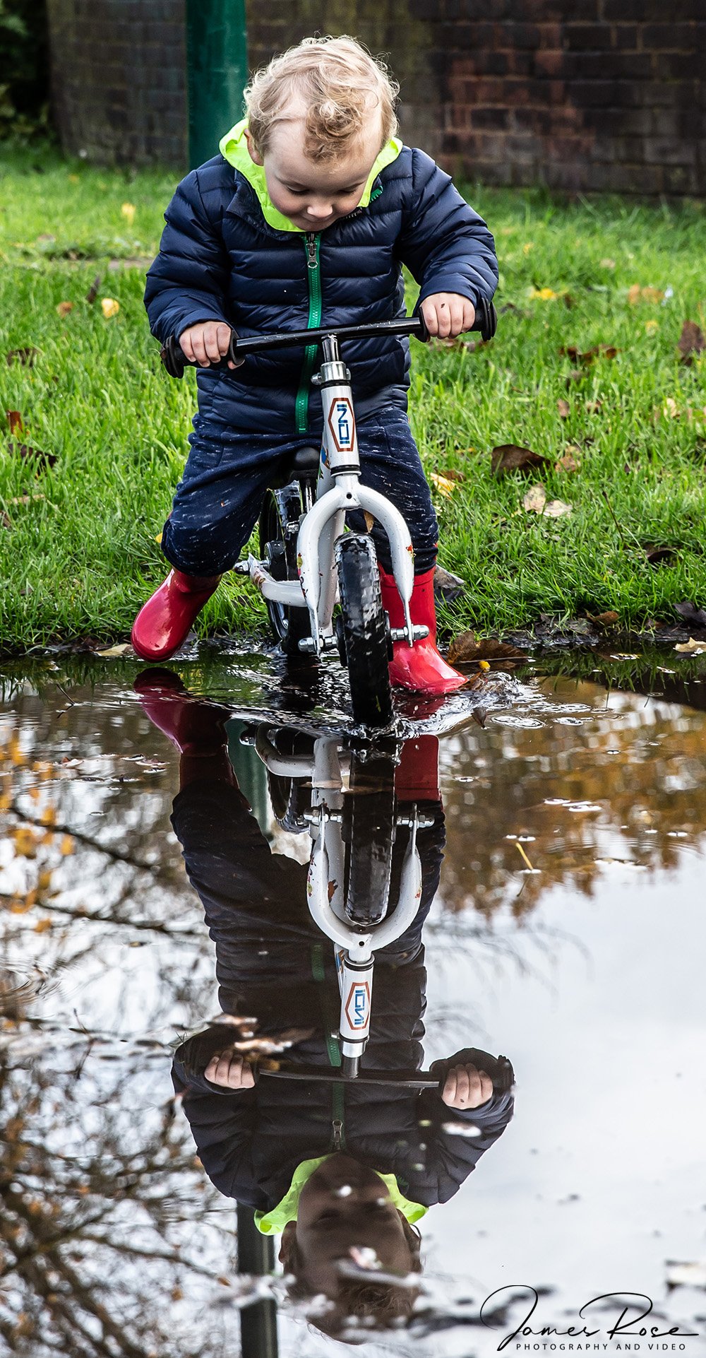 A young child riding a bicycle through a puddle on a grassy area, with reflection visible in the water.