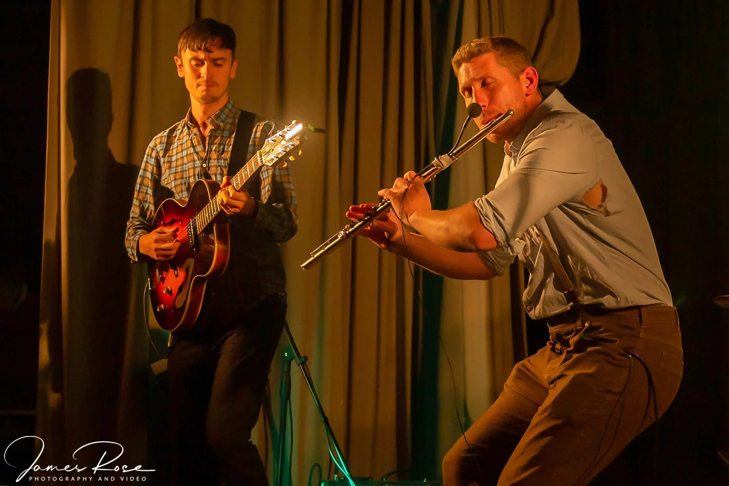 Two musicians performing on a stage. One is playing an acoustic guitar, the other is playing a flute. They are illuminated by warm stage lighting with curtains in the background.