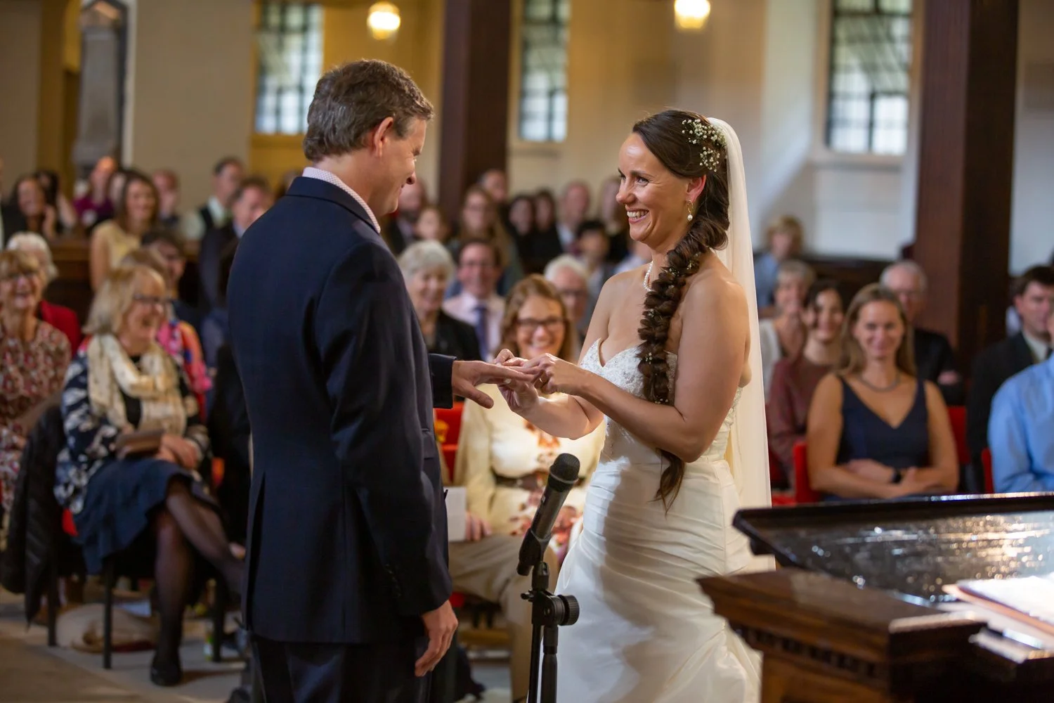 A bride and groom exchanging wedding vows in a church, smiling at each other while holding hands, with guests seated in the background.