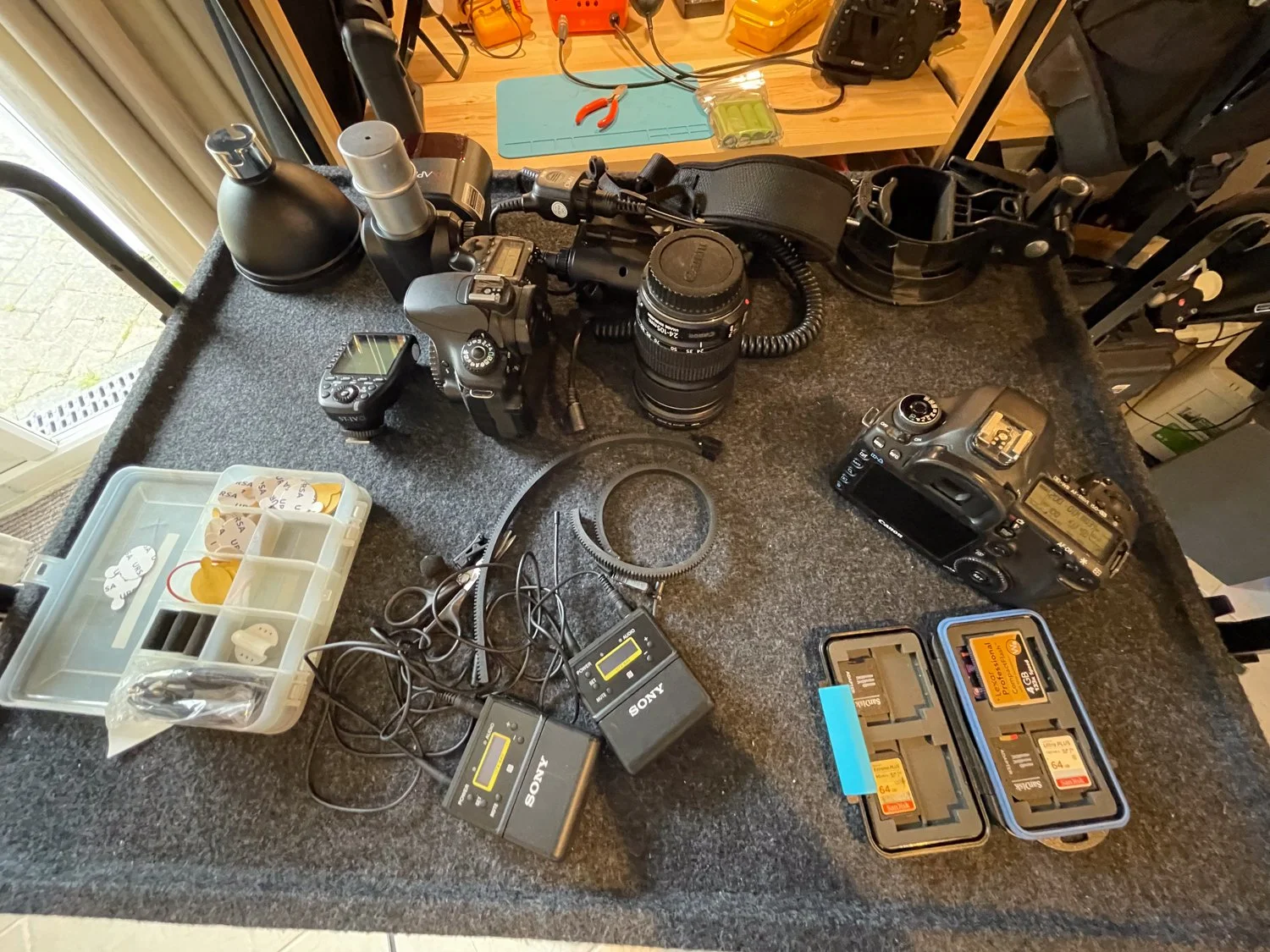 A photography workspace with two digital cameras, one with a lens detached, two battery chargers, camera batteries, a small plastic container with camera accessories, a set of small tools like pliers and wire cutters, and various camera gear on a dar