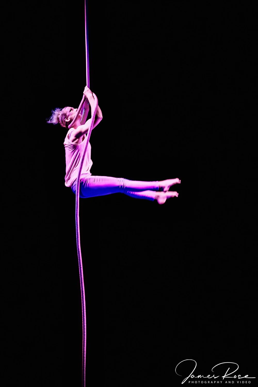 A female aerial acrobat performing on a pink silk fabric against a black background, wearing a light pink top and blue leggings.