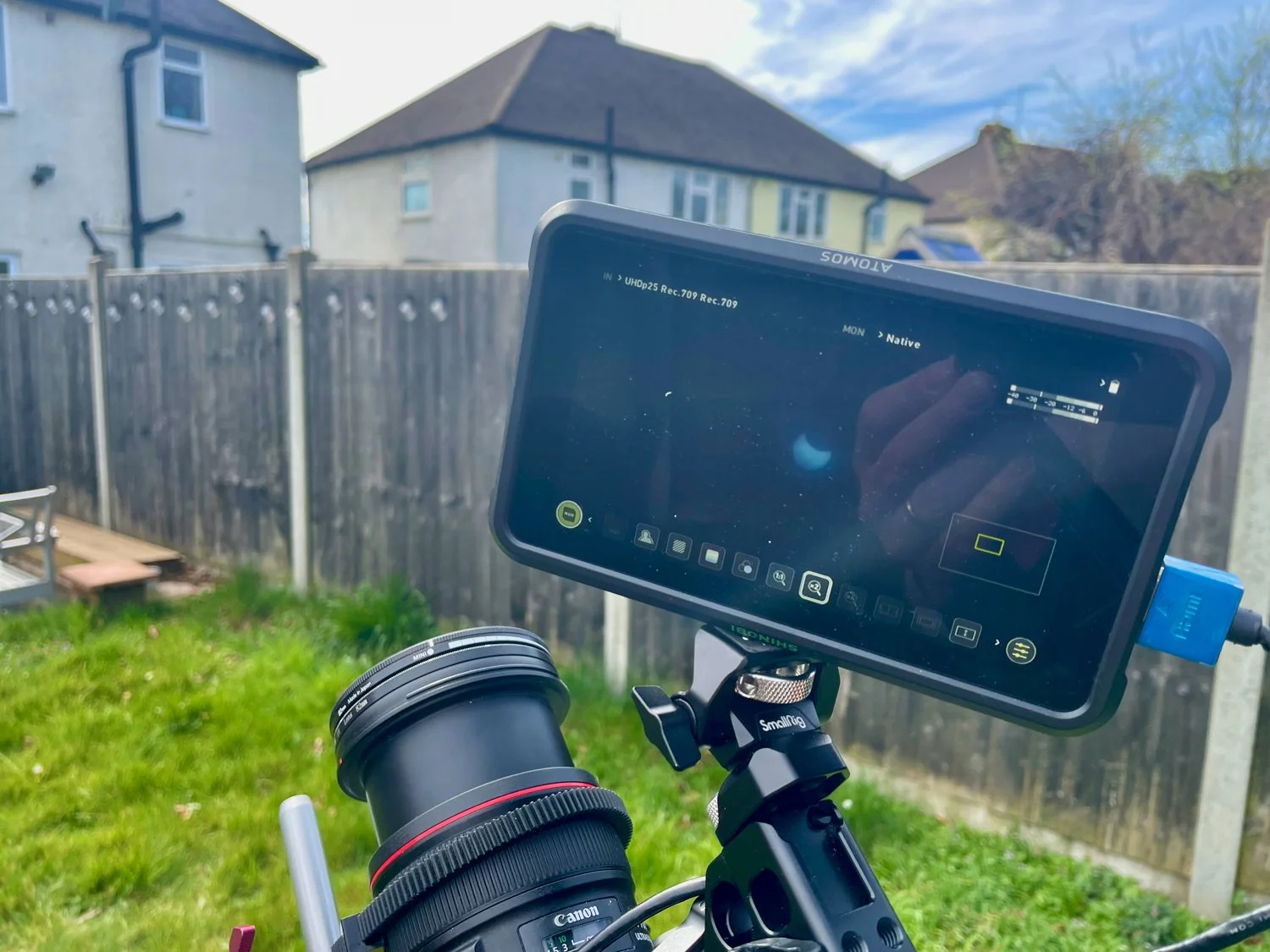 A camera rig with a Canon camera and a monitor displaying a view of the moon in a backyard with a wooden fence, green grass, and neighboring houses under a blue sky.