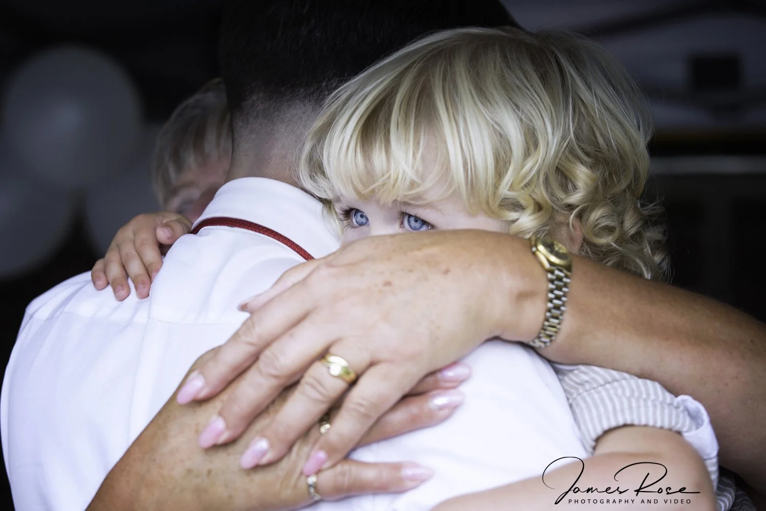 A young girl with blonde curly hair and blue eyes hugging an adult, possibly her father, with only the back of his head visible and a glimpse of another child in the background.