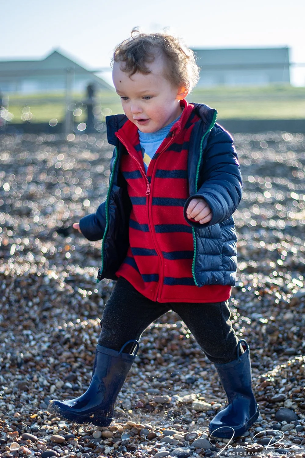 A young boy in a blue puffy jacket, red hoodie, and rain boots walking on a rocky surface outdoors, with houses and a fence in the background.