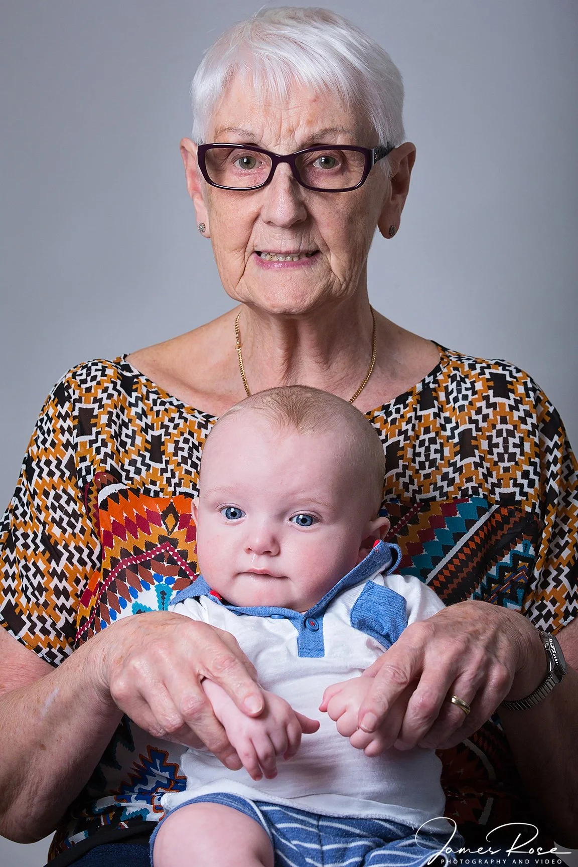 An elderly woman with white hair and glasses holding a baby with blue eyes and light hair, both facing forward against a plain gray background.