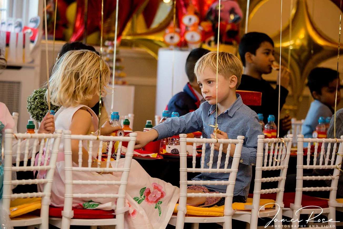 Children sitting at a decorated party table, with a boy handing a snack to a girl, under hanging balloons and party decorations.