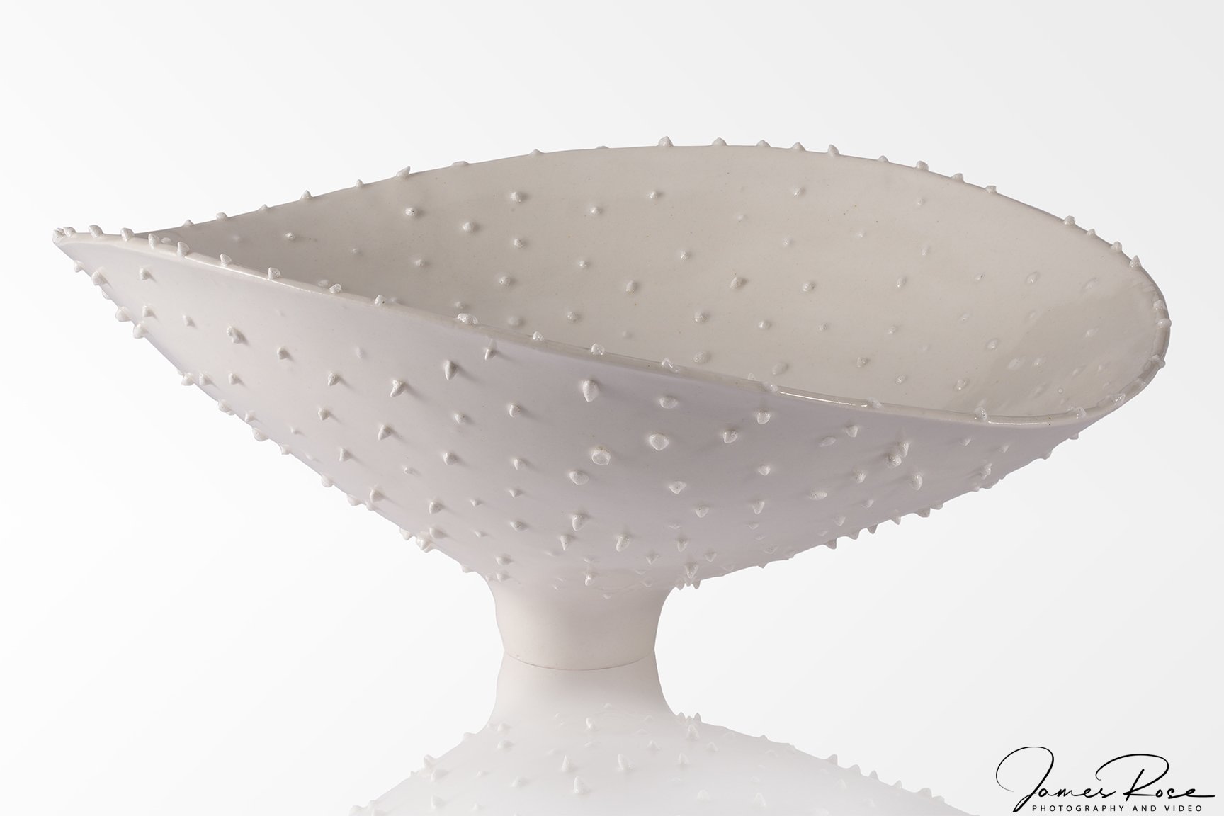A white ceramic bowl with a textured surface covered in raised bumps, placed on a reflective surface against a plain white background.