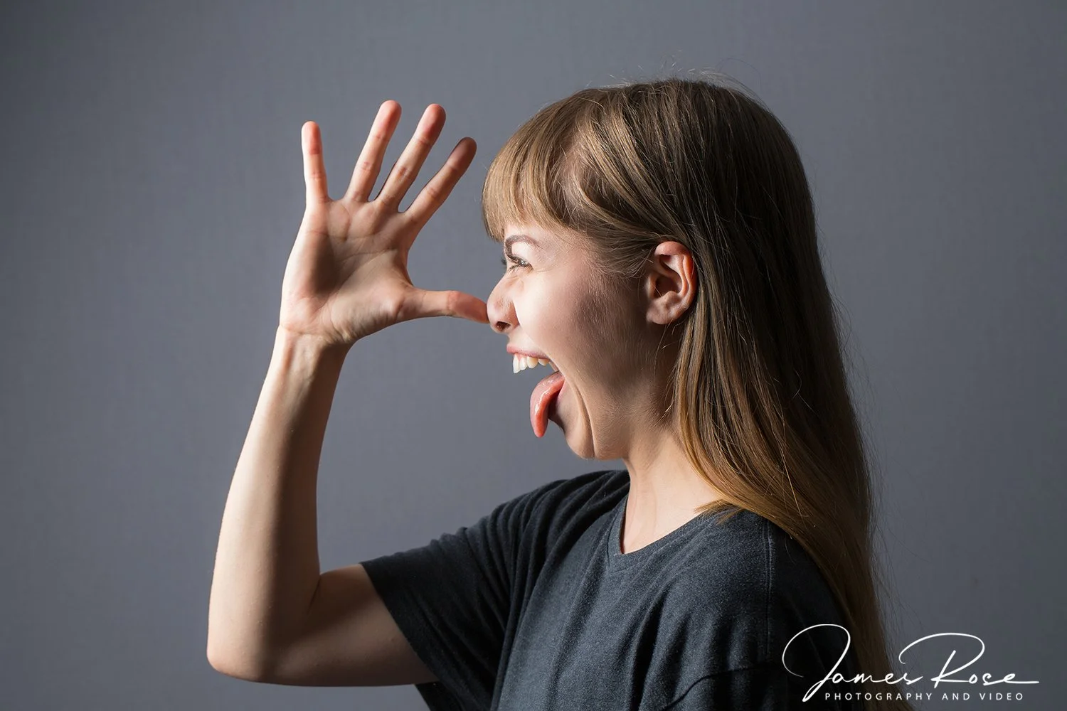 Young woman with light brown hair making a funny face with her tongue out and hand near her face against a gray background.
