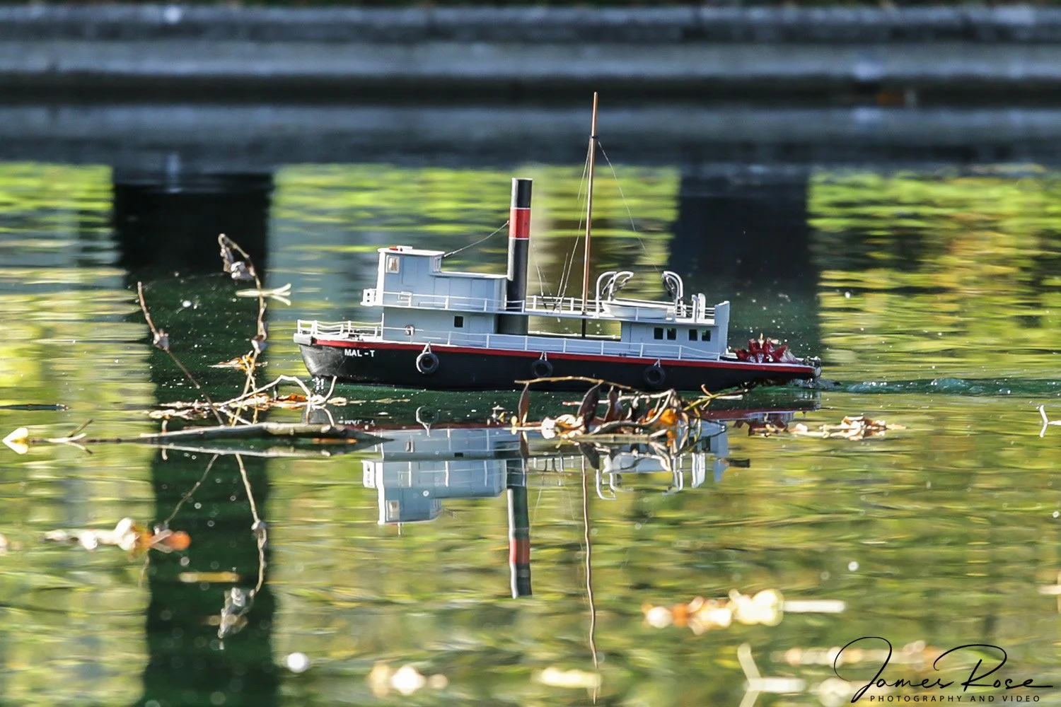 A small model boat floats on a body of water with floating debris and reflections of trees and structures in the background.