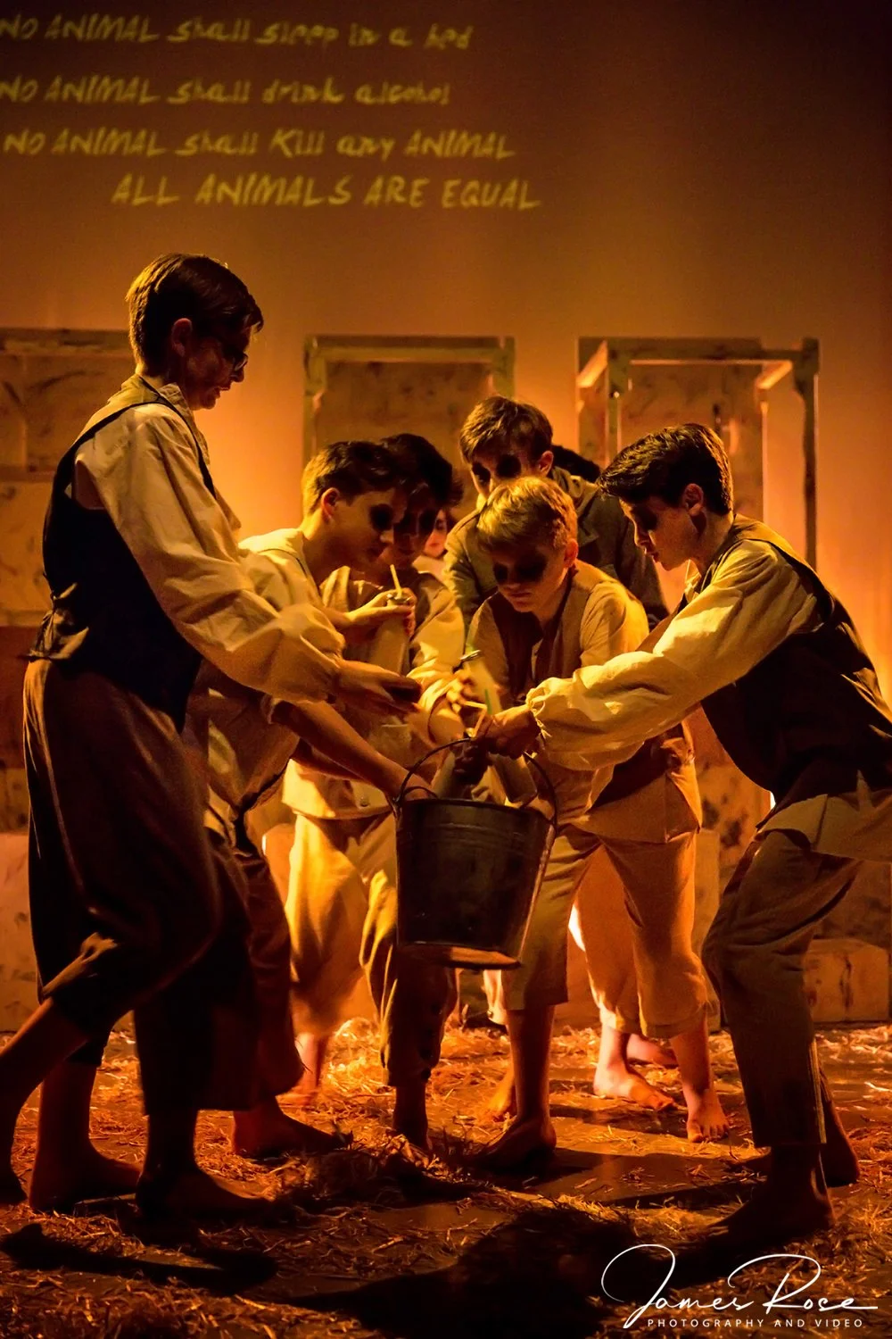 A group of children dressed in old-fashioned clothing, holding a bucket and trying to catch or capture something on a stage with wooden crates and a warm, gold-toned lighting. A quote about animal cruelty is projected on the wall in the background.