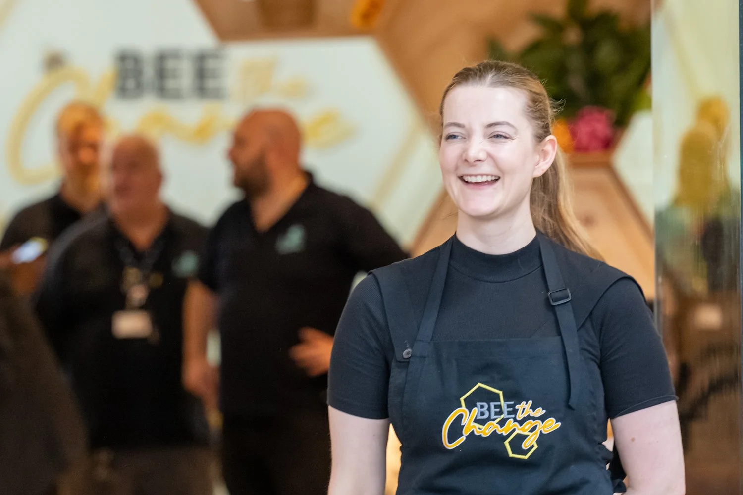 A smiling woman wearing a black t-shirt and apron with a logo reading 'BEE the Change' is in the foreground. In the background, three people are talking, slightly out of focus, in an indoor setting with a sign that also says 'BEE' and some decorative