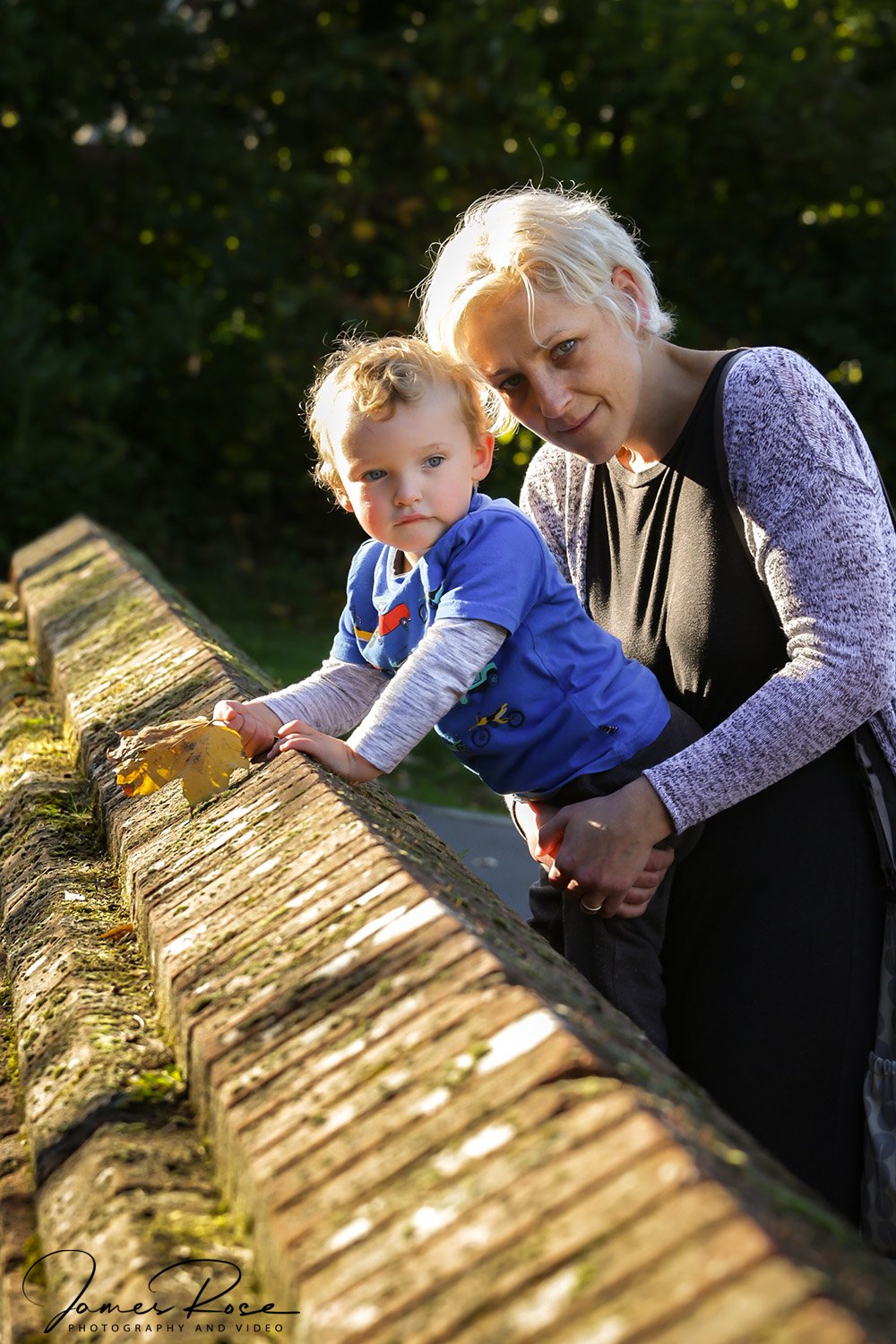 A woman and a young boy leaning over a moss-covered brick ledge outdoors, with the woman holding the boy as he reaches for a yellow leaf. They are in a sunlit green area.