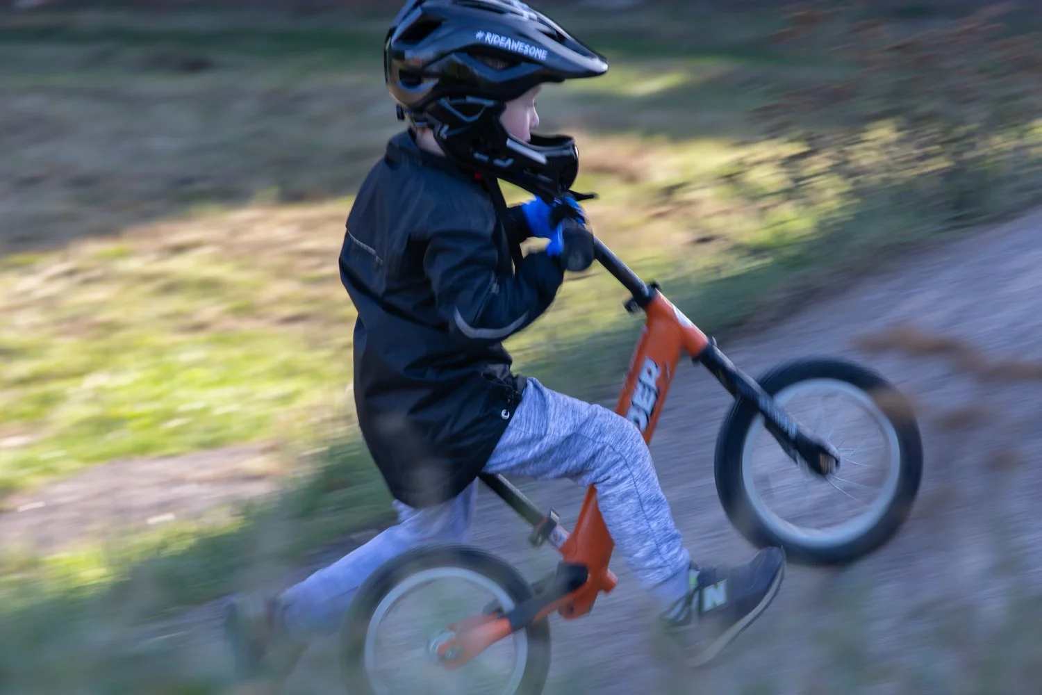 A young child wearing a black helmet and jacket riding an orange balance bike on a dirt trail with green grass and trees in the background.