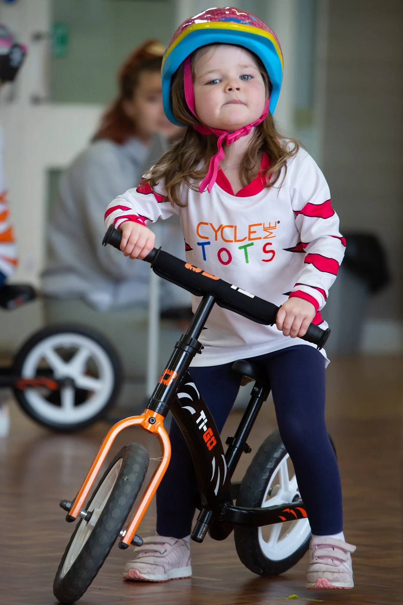 Young girl wearing a colorful helmet riding a black and orange balance bike indoors. She has long brown hair, is dressed in a white shirt with red accents and the words 'Cycle Me Tots' on it, and wears navy blue pants and pink shoes.