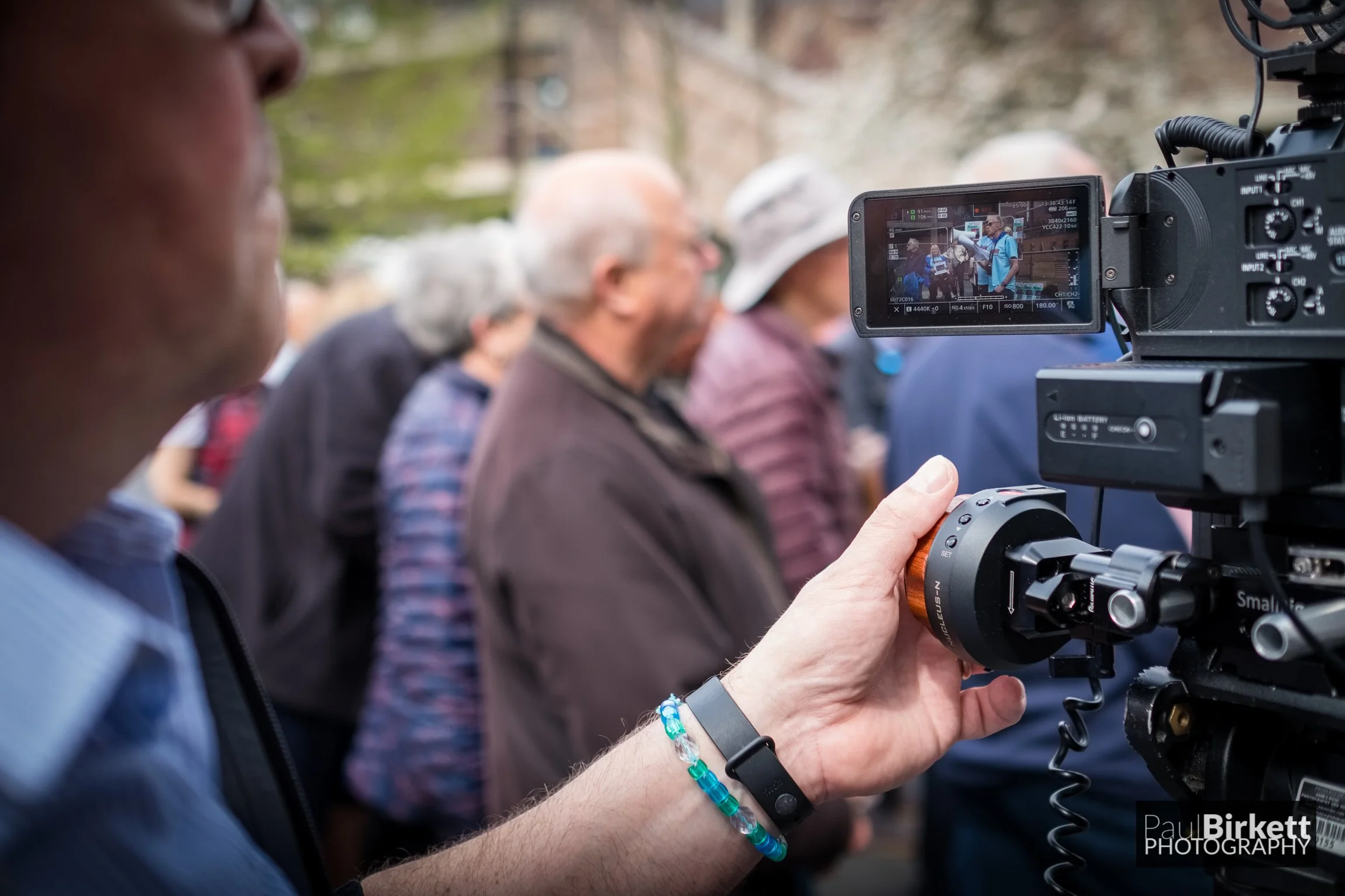 A person operating a professional video camera during an outdoor event with a crowd of people in the background.