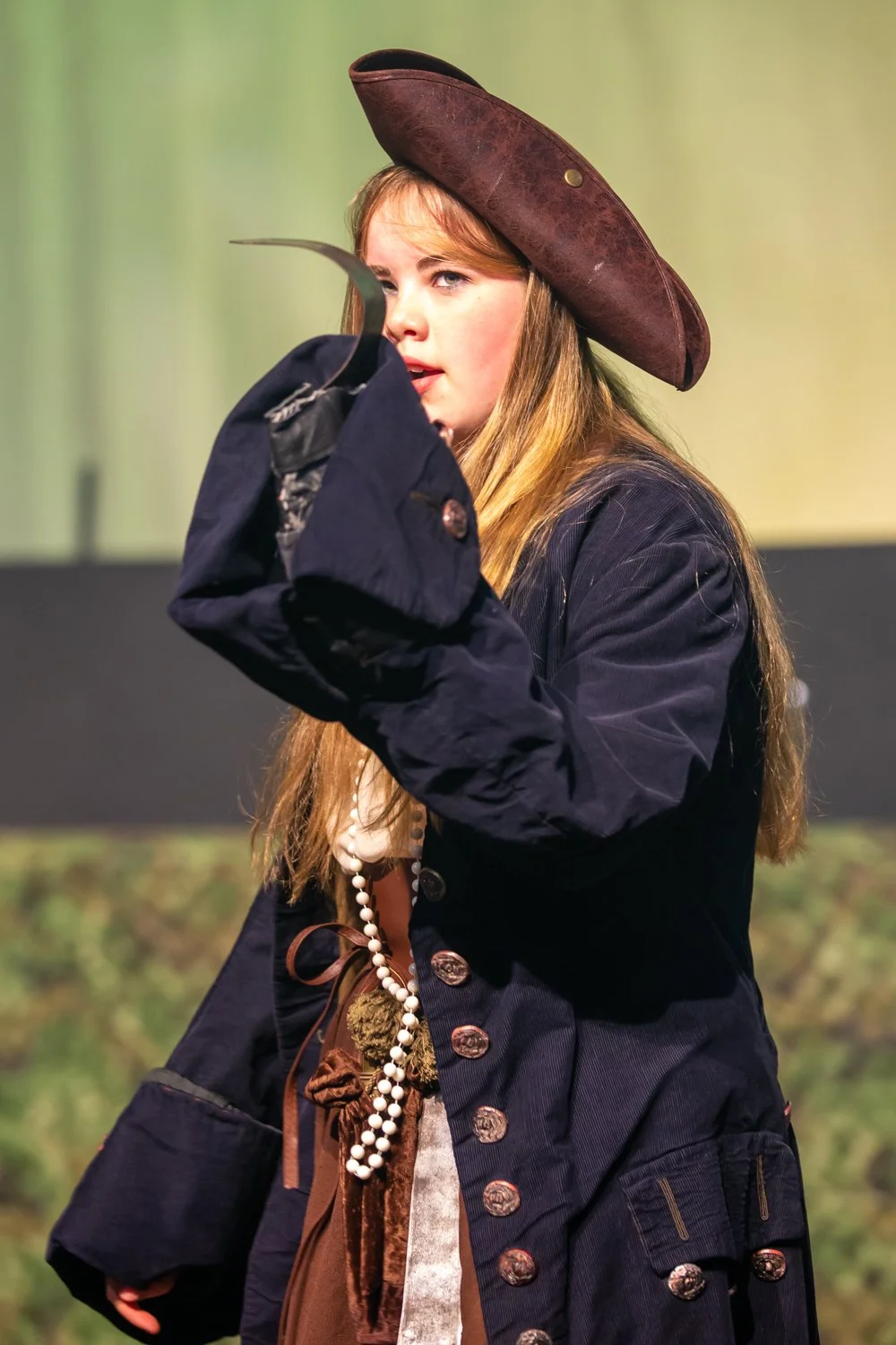 A young woman dressed as pirates, wearing a large brown felt hat, a navy blue coat, and holding a hook in front of her mouth, appears to be performing or acting on stage.