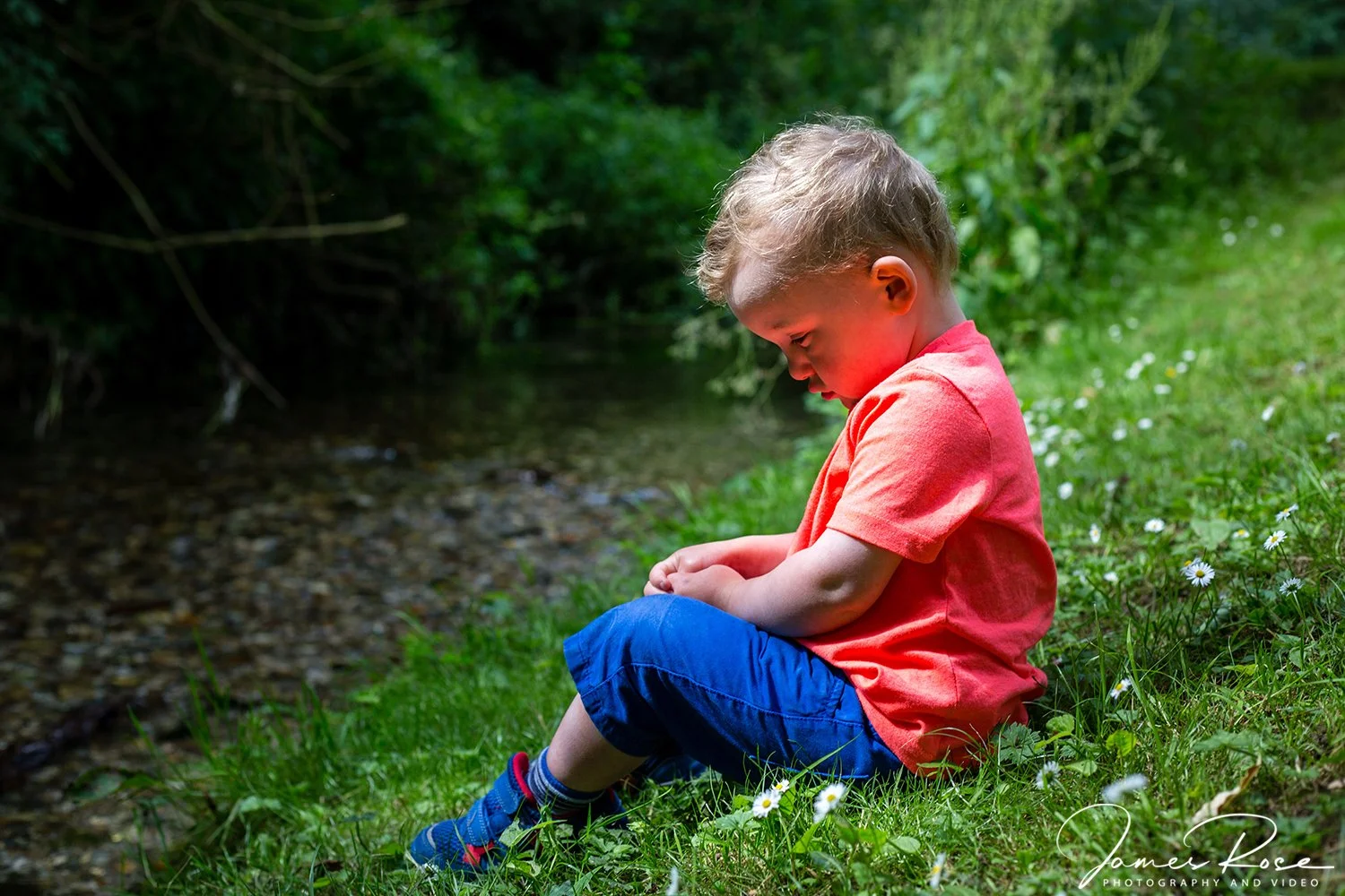 Young boy sitting on grass near a stream, looking down at his lap, surrounded by greenery and small white flowers.
