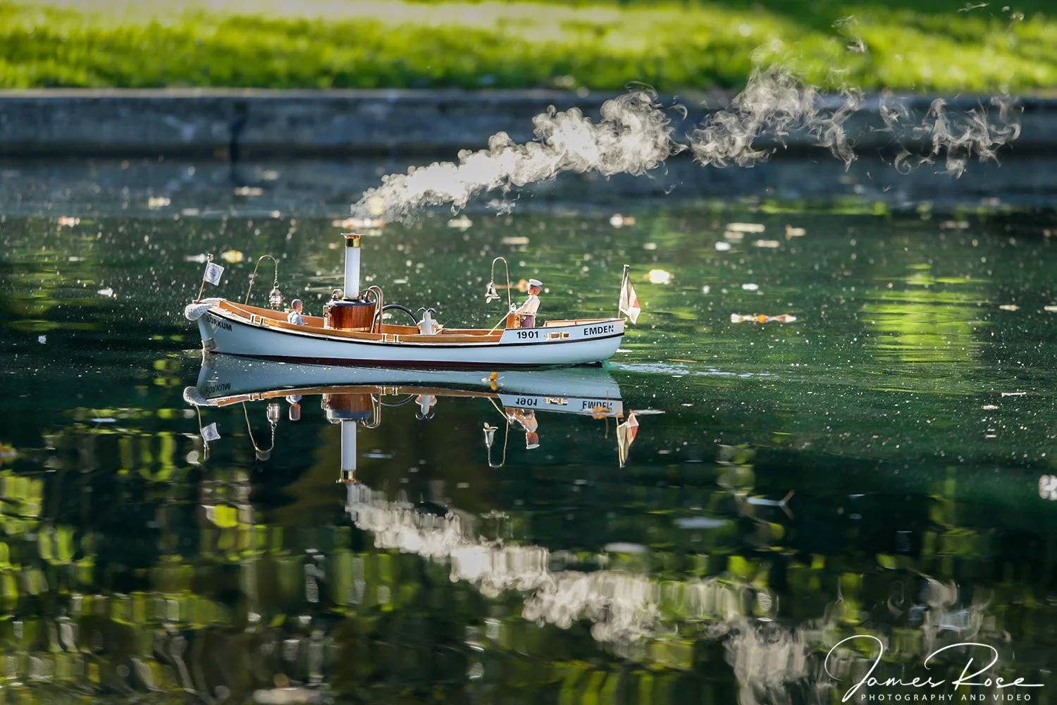 A detailed model of a vintage steam-powered boat on a calm lake, with a wooden cabin, a small chimney emitting smoke, and tiny human figures on board, reflected in the water with green foliage in the background.
