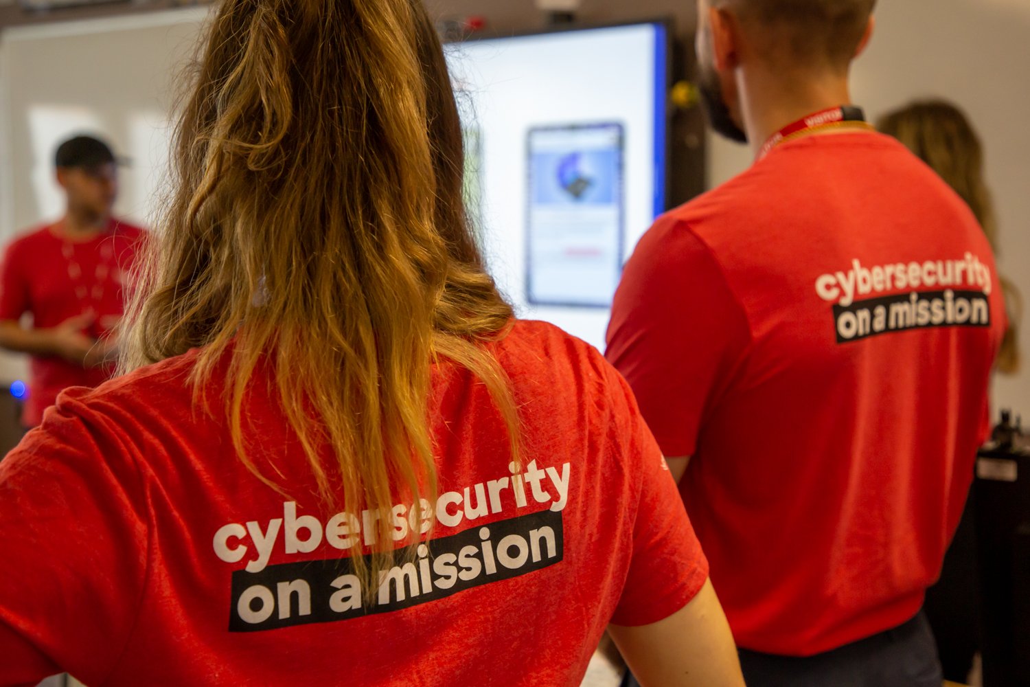 People wearing red t-shirts with the text 'cybersecurity on a mission', working together in front of a computer screen.
