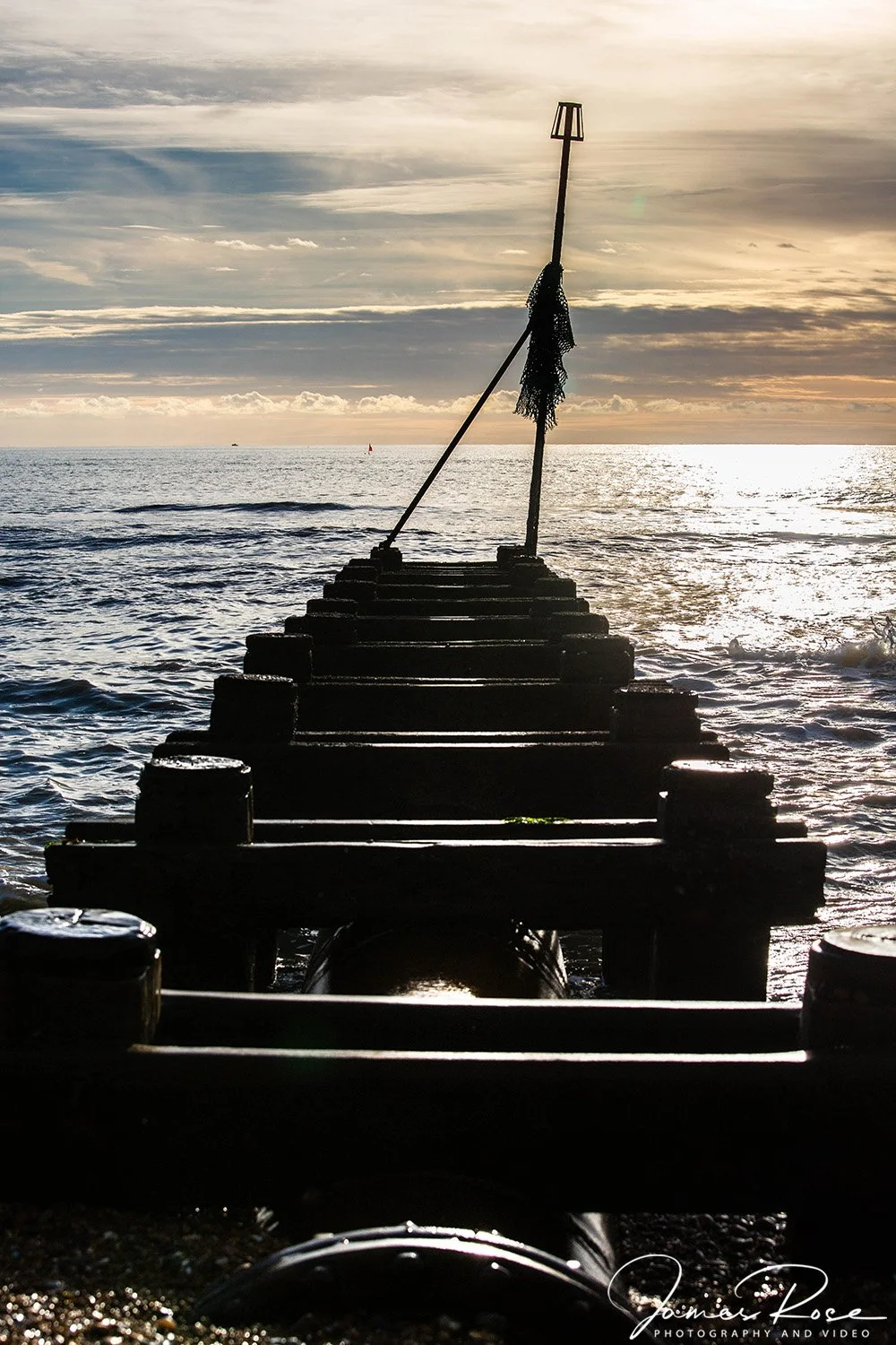 Silhouette of a wooden pier extending into the ocean during sunset with a flagpole at the end of the pier, the sky filled with clouds and the sun reflecting on the water.