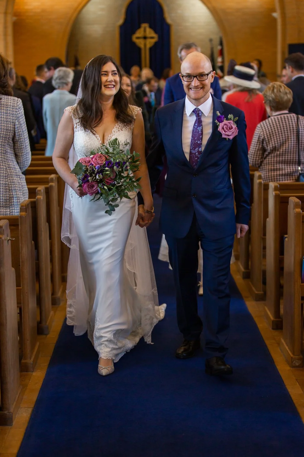 A bride and groom walking down the aisle inside a church, smiling and holding hands, with the bride holding a bouquet of purple and pink flowers.