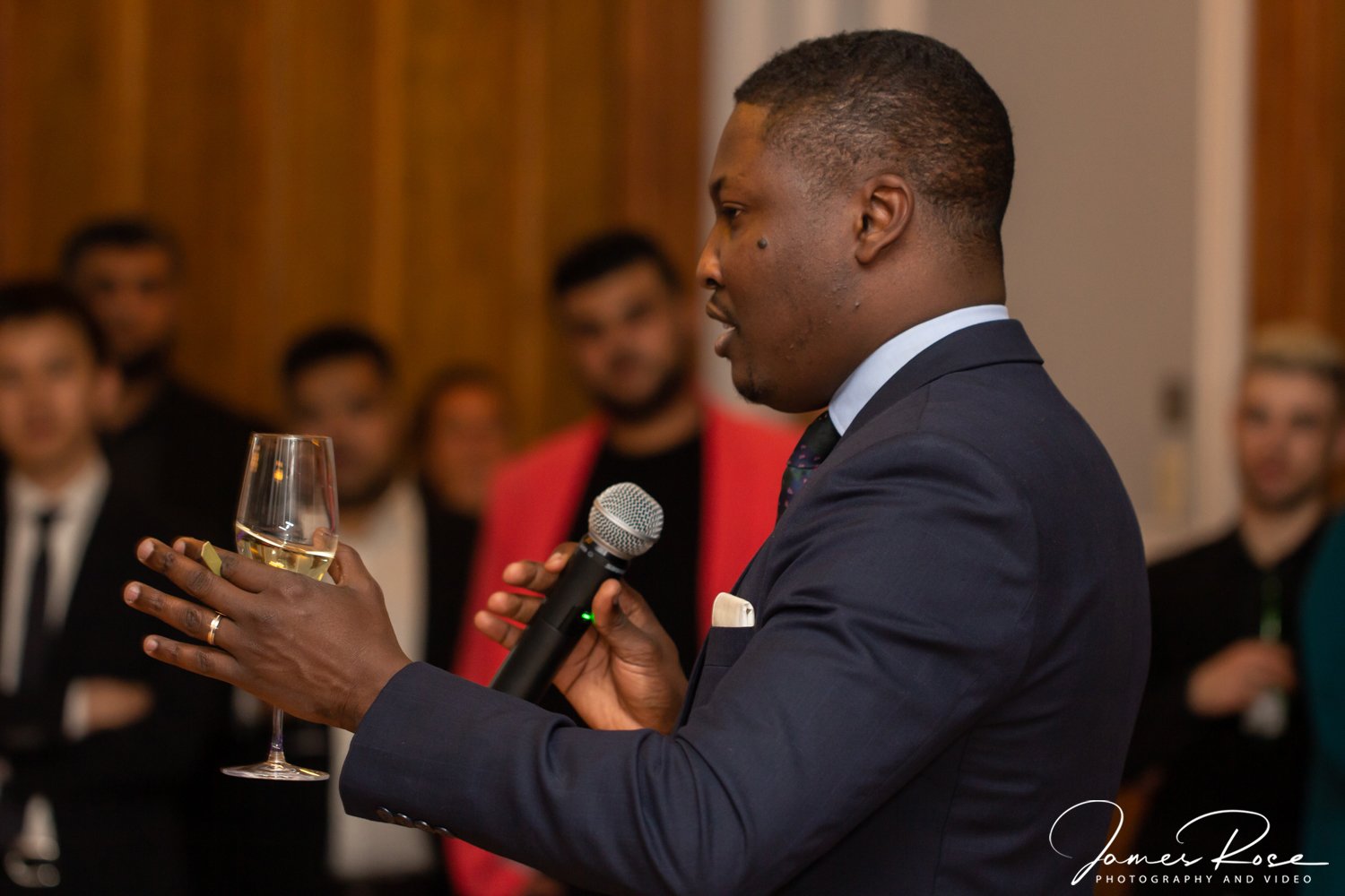 A man in a dark suit holding a wine glass in one hand and a microphone in the other, speaking at an event with a blurred group of people in the background.