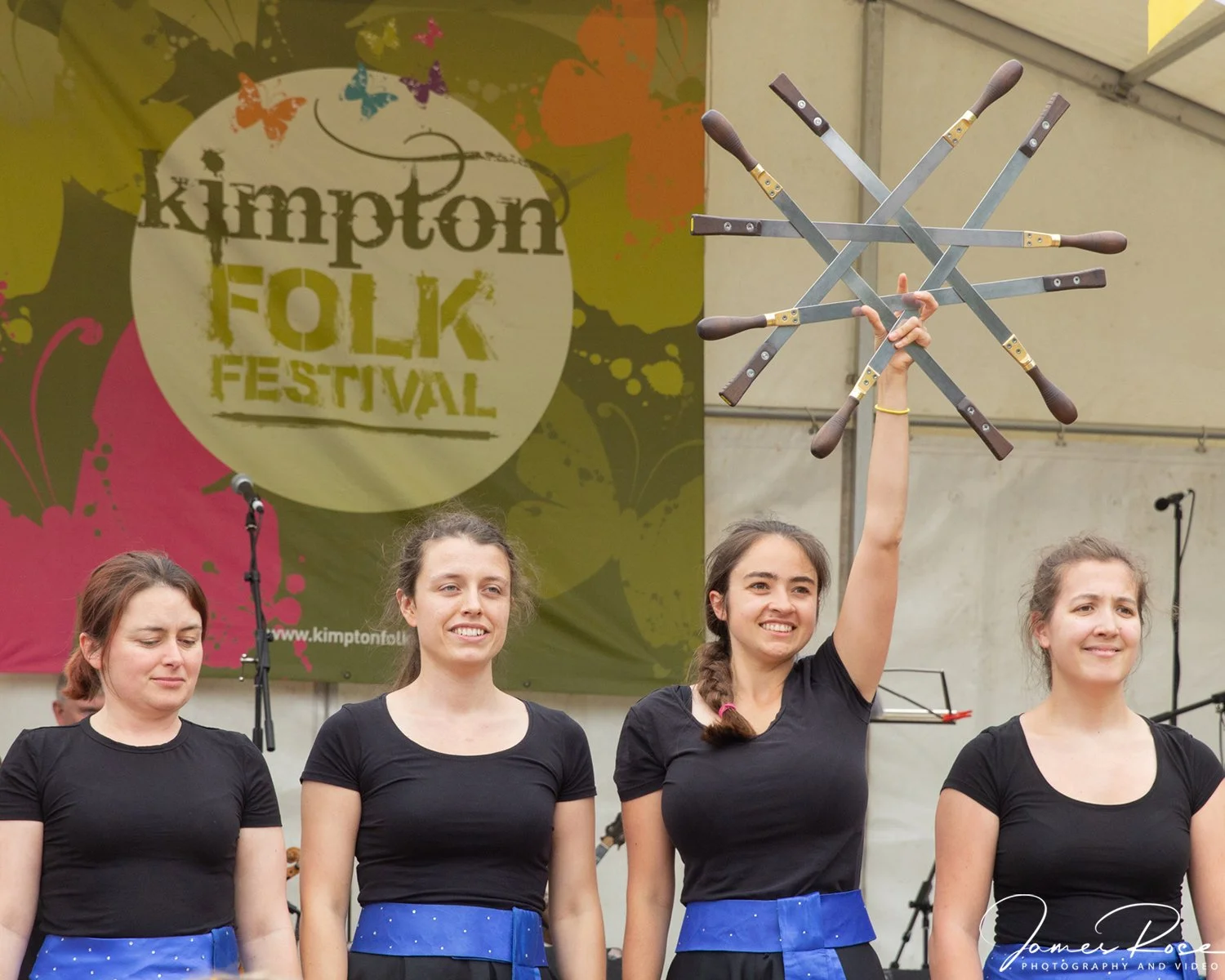 Four women in black shirts and blue skirts standing on stage at the Kimpton Folk Festival, one woman is holding multiple folding knives in the air.