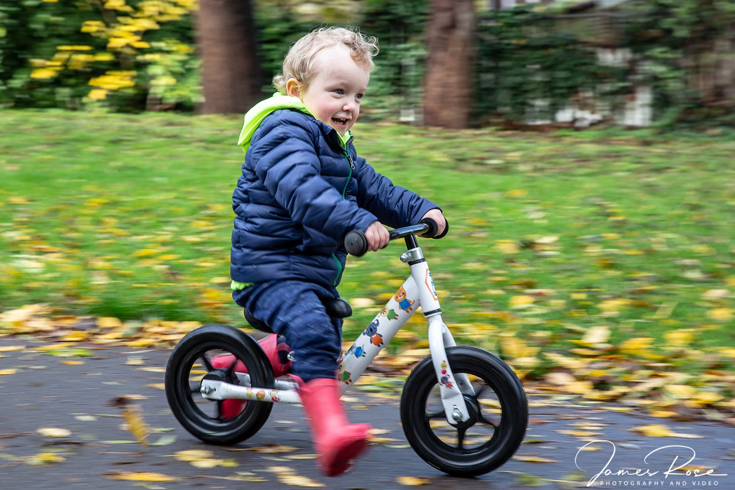 A young boy wearing a blue jacket and pink rain boots riding a white bicycle with colorful animal stickers through a park with yellow fallen leaves and green grass.