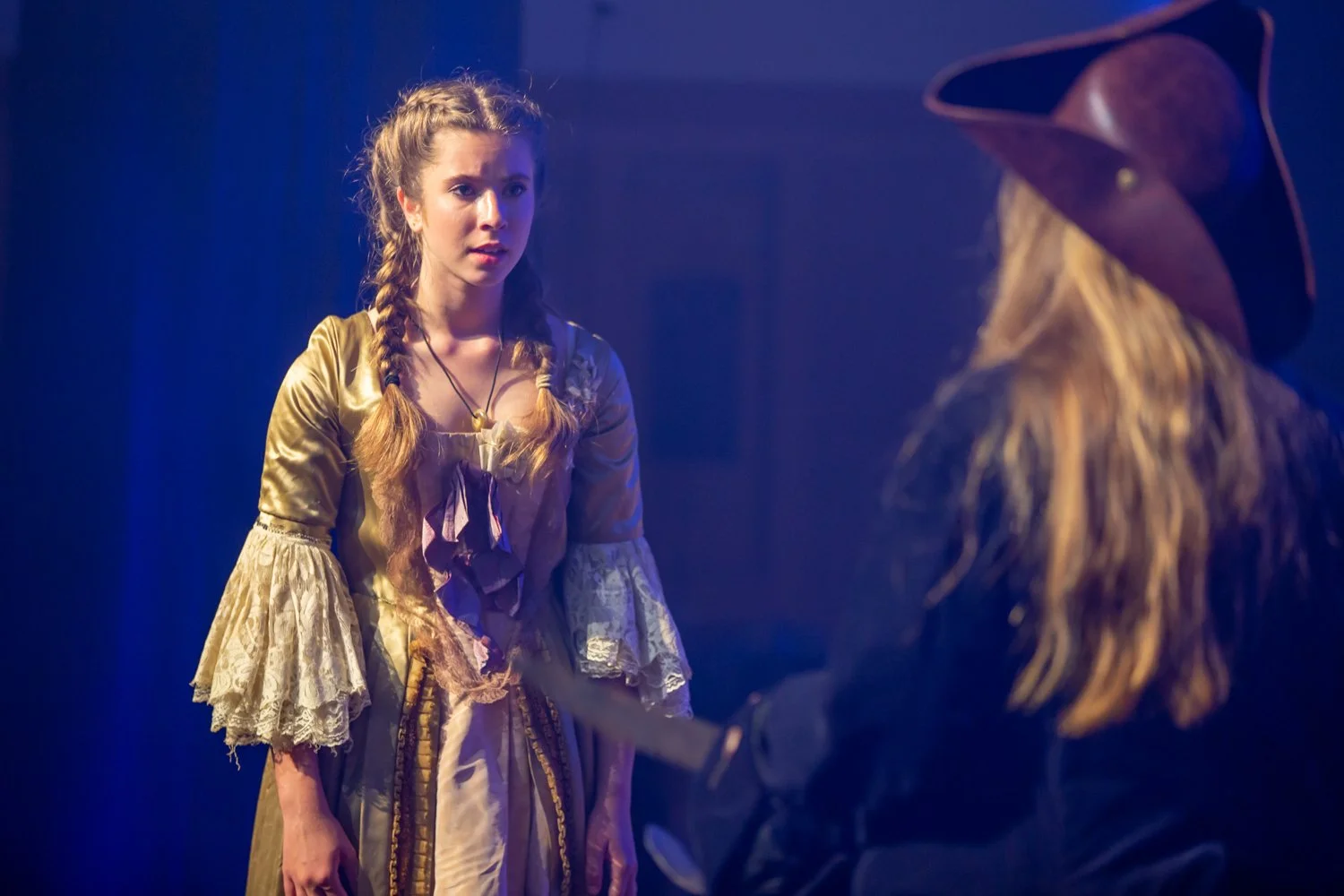 Two women in costumes, one with braids and a vintage dress, and another with a witch hat and dark clothing, facing each other during a theatrical performance.