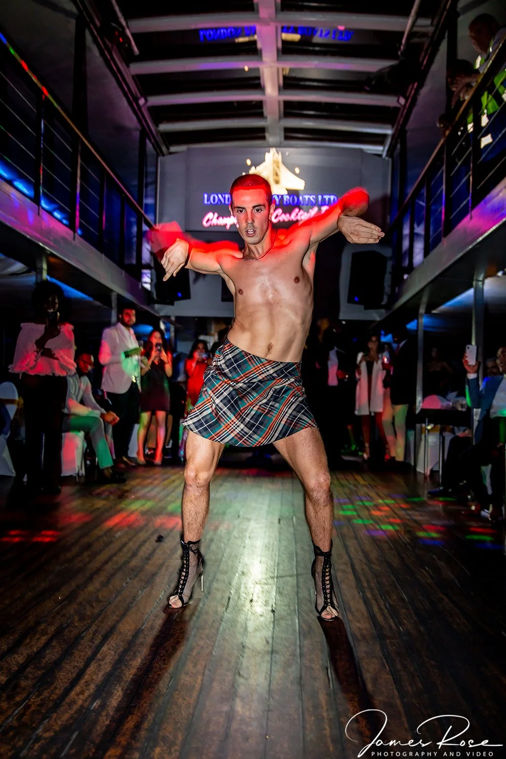 A shirtless man with red hair posing in a kilt and high heels at a lively nightclub or party with people watching in the background.