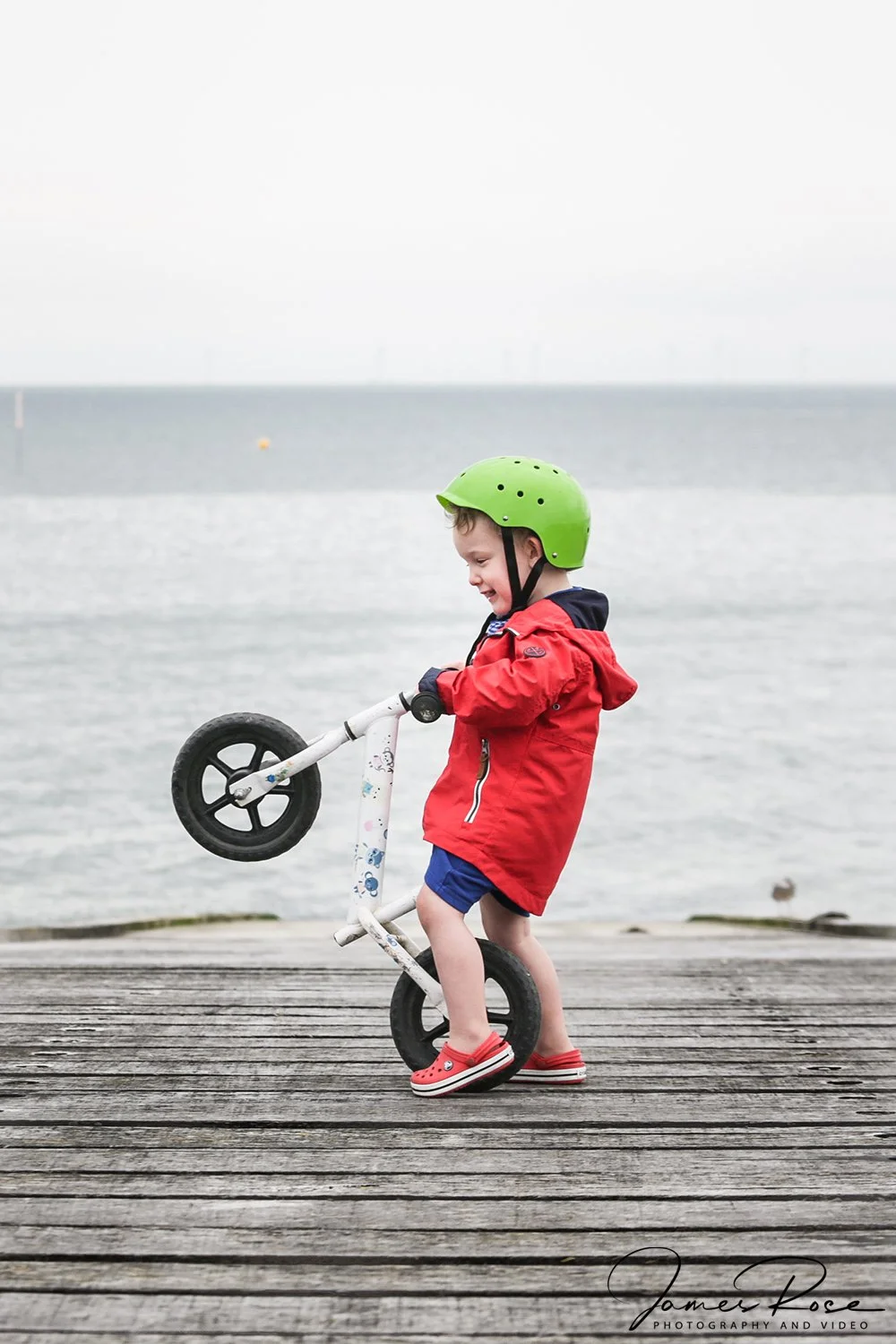 A young boy wearing a green helmet, red jacket, blue shorts, and red sneakers standing on a wooden dock near the water, lifting his white balance bike with black wheels.