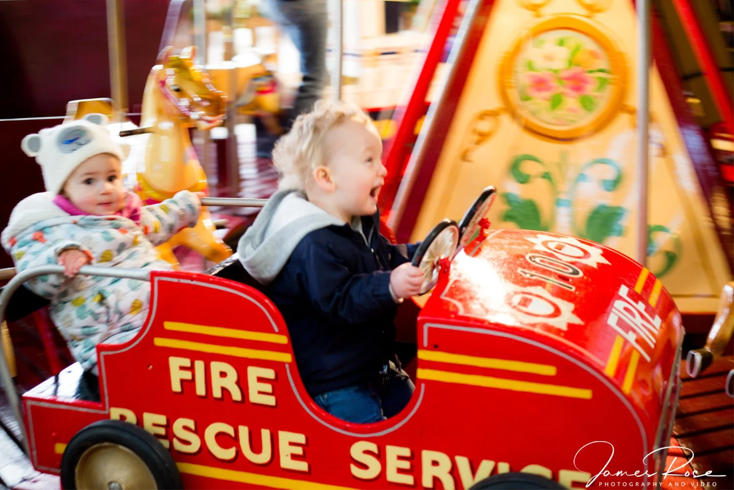 Two children riding in a colorful amusement park fire truck carousel. The boy, in a dark jacket, is smiling and holding the steering wheel. The girl, wearing a bear hat and a colorful coat, looks toward the camera. The carousel is bright red with yel