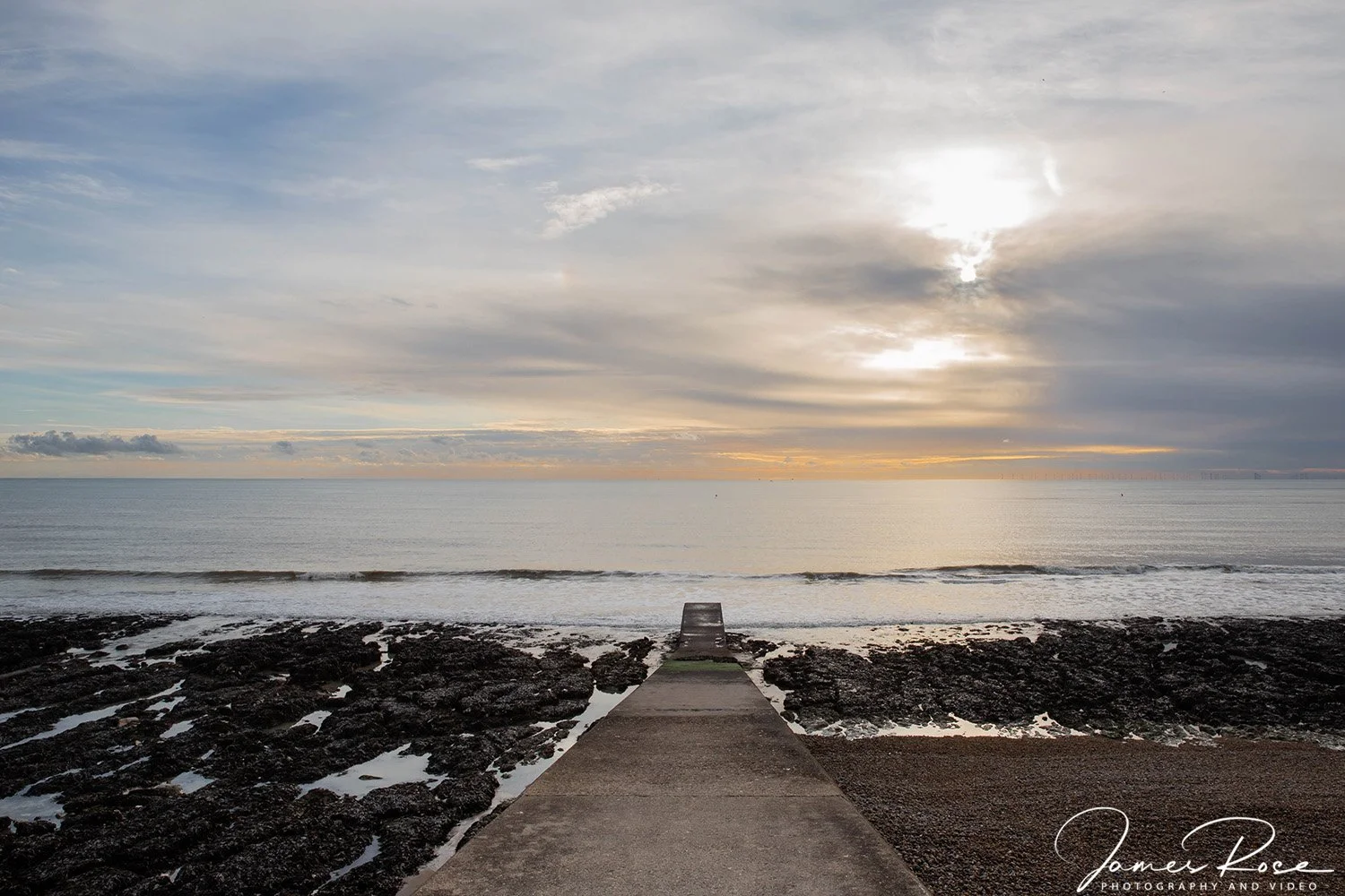 A concrete pier leading to a calm ocean during a cloudy sunset, with a cloudy sky and the sun partially visible, casting a soft light.