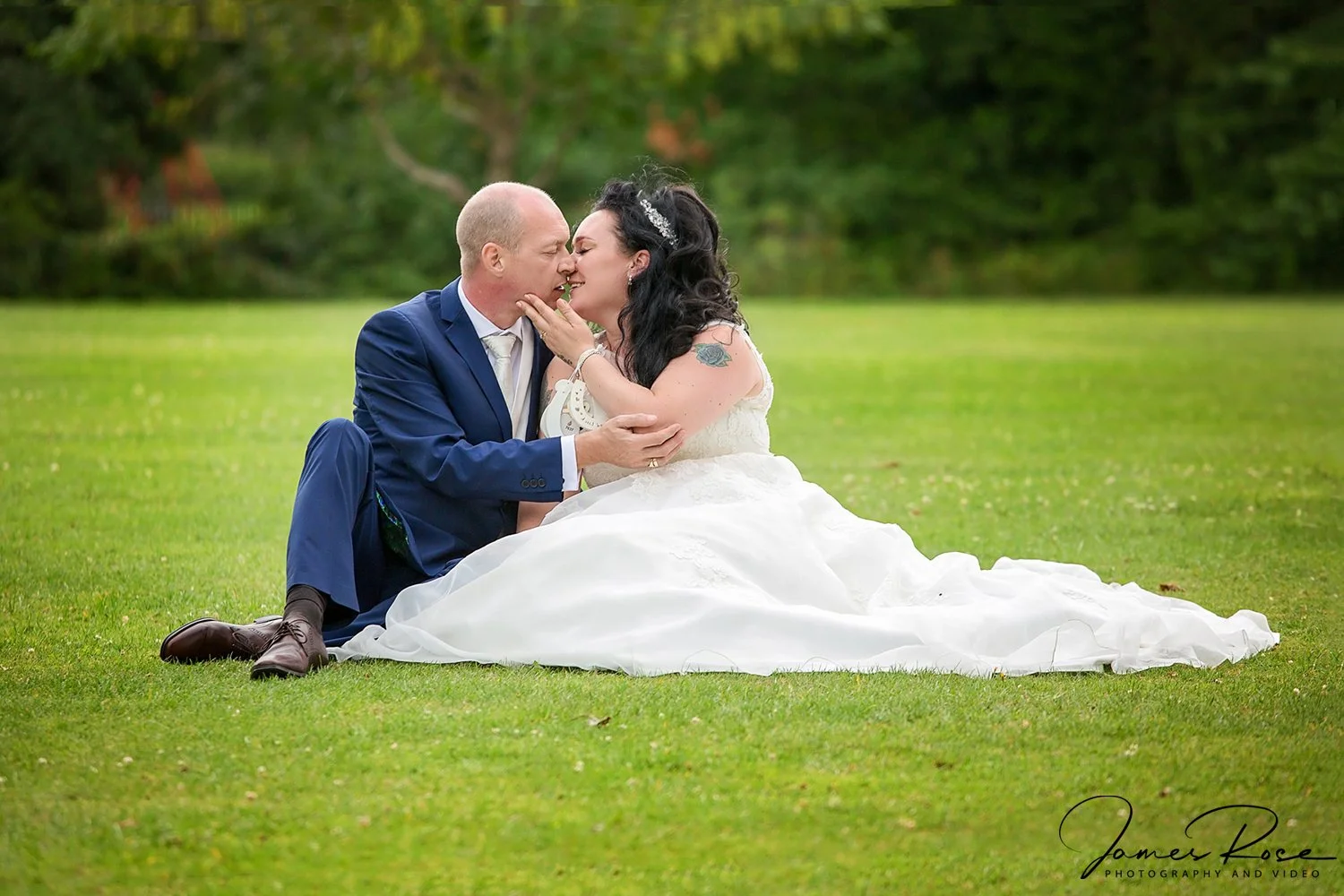 A newlywed couple sitting on the grass, the groom in a blue suit and the bride in a white wedding dress, sharing a tender moment outdoors.