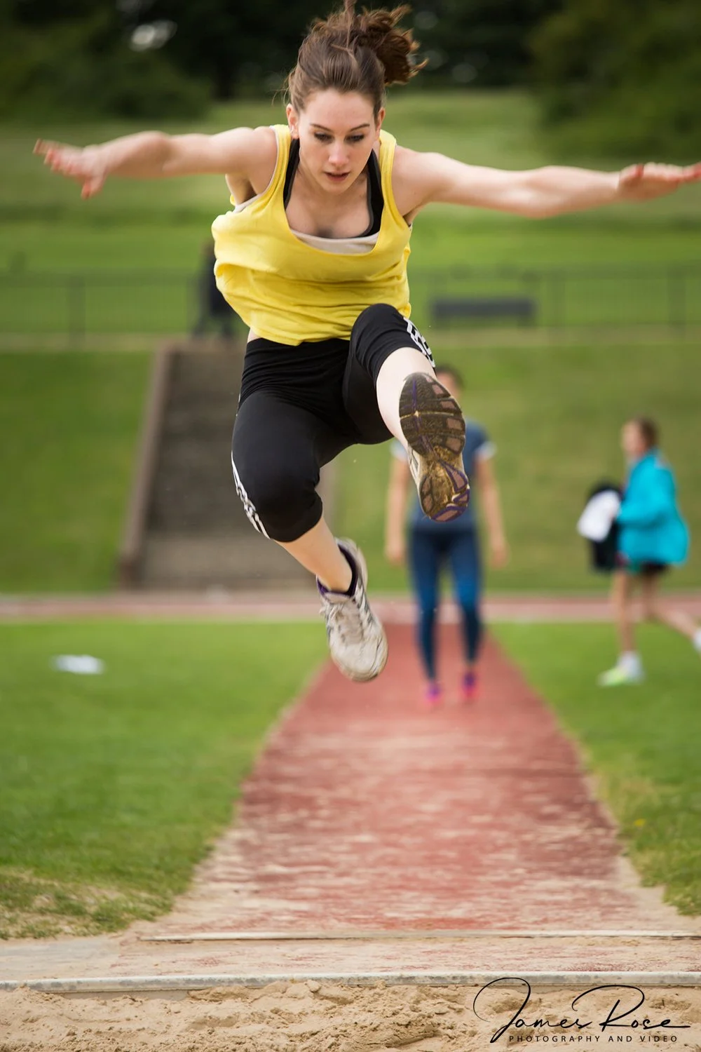 Female athlete in yellow tank top and black shorts jumping into a sandpit during a track and field event.
