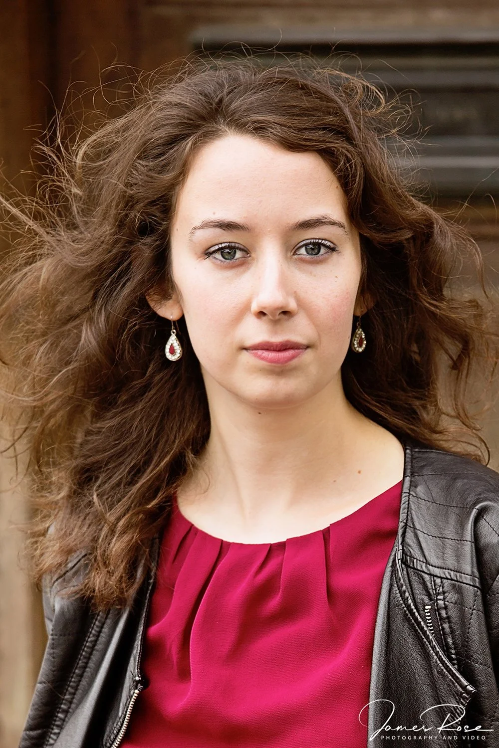 Portrait of a young woman with curly brown hair, light green eyes, wearing earrings, a maroon blouse, and a black leather jacket, standing outdoors with a blurred wooden background.