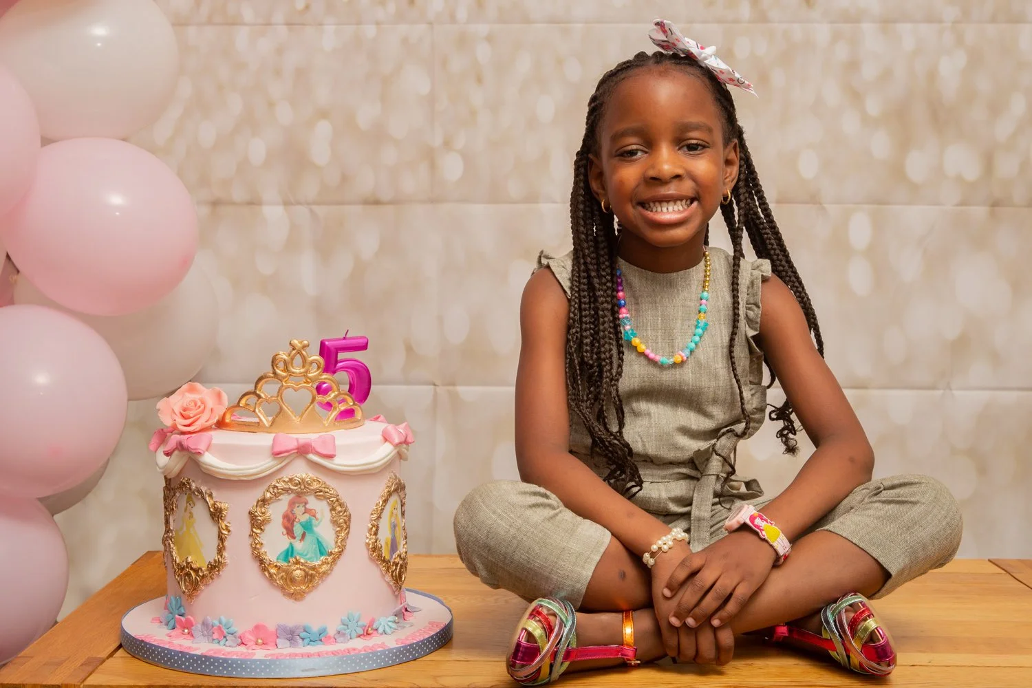 A young girl sitting on a wooden table with a pink princess-themed birthday cake and pink balloons to her left. The girl has braided hair, wears colorful jewelry, a green outfit, and rainbow-colored shoes, and is smiling.