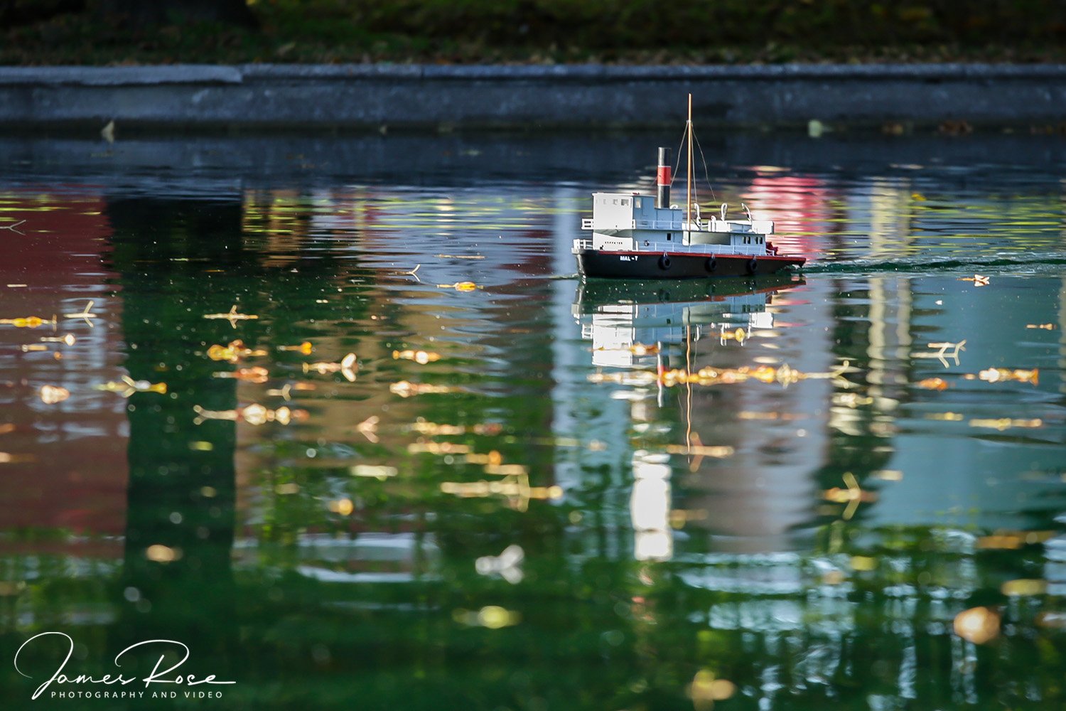 A small model boat floating on water with autumn leaves, colorful reflections, and a blurred background.