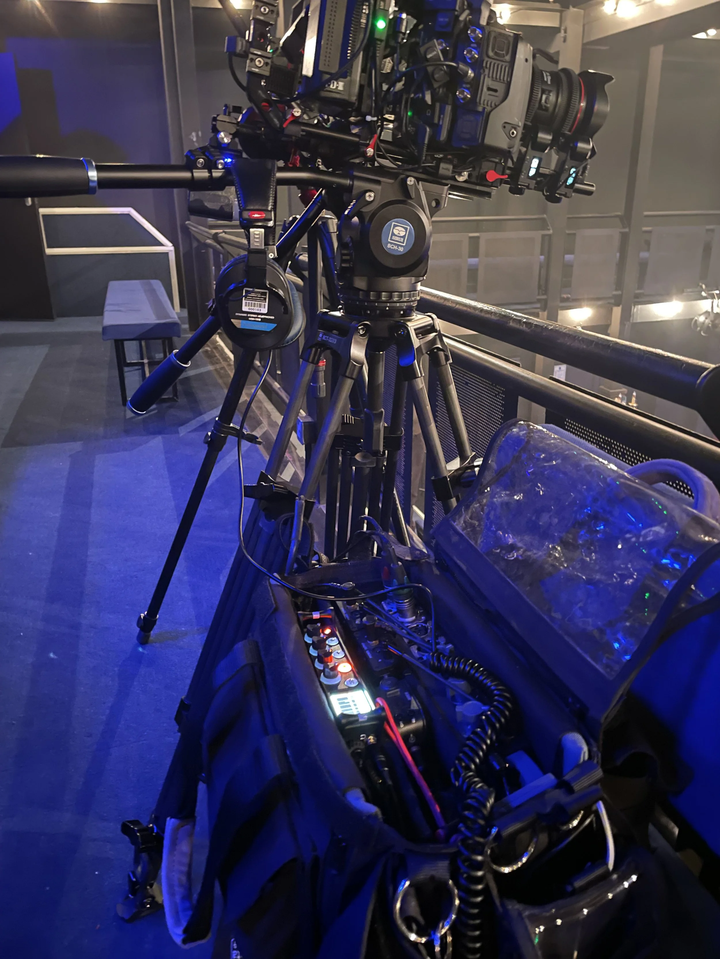 A professional film camera mounted on a tripod, with various filming equipment and controls visible, set in a dark, industrial-looking studio or stage environment.