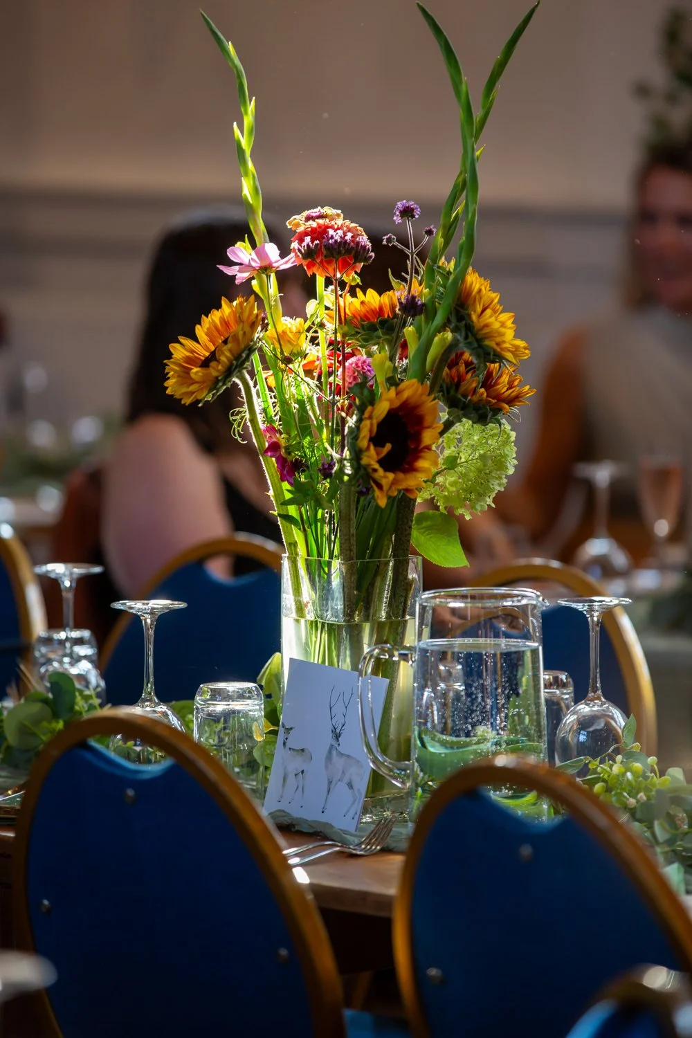 A tall glass vase with colorful flowers including sunflowers and various other blooms, placed on a table set with glassware and a card with deer illustration, at an indoor event.