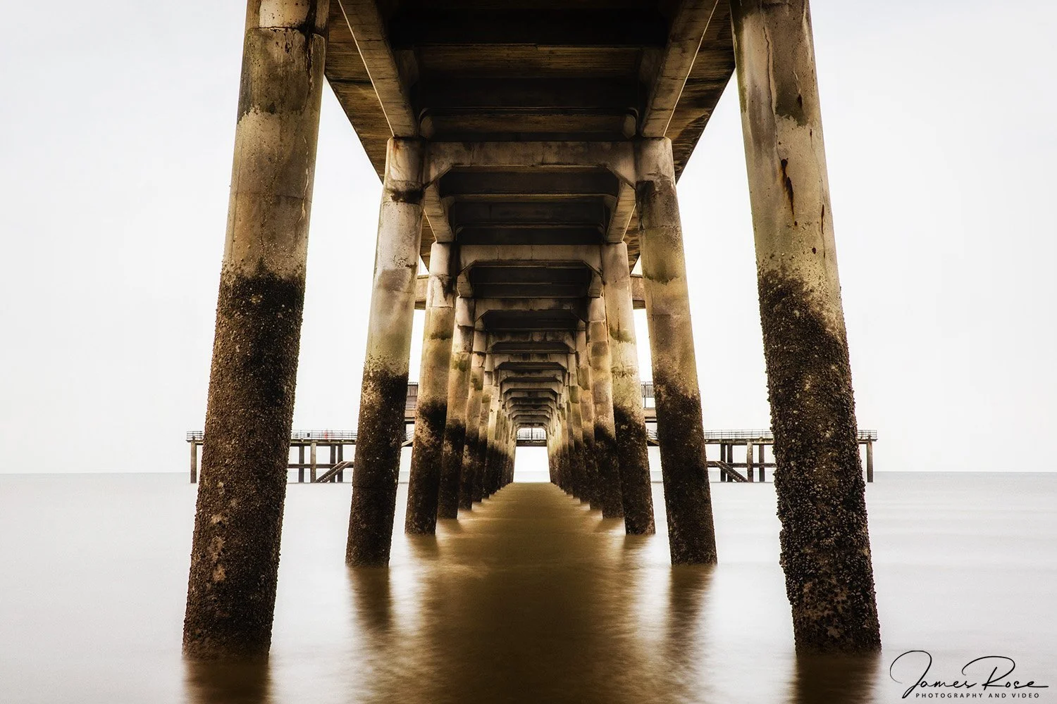 Looking up from under a pier over the ocean, showing the concrete and steel structure of the pier's supports extending into the water with a small distant frame of the horizon.
