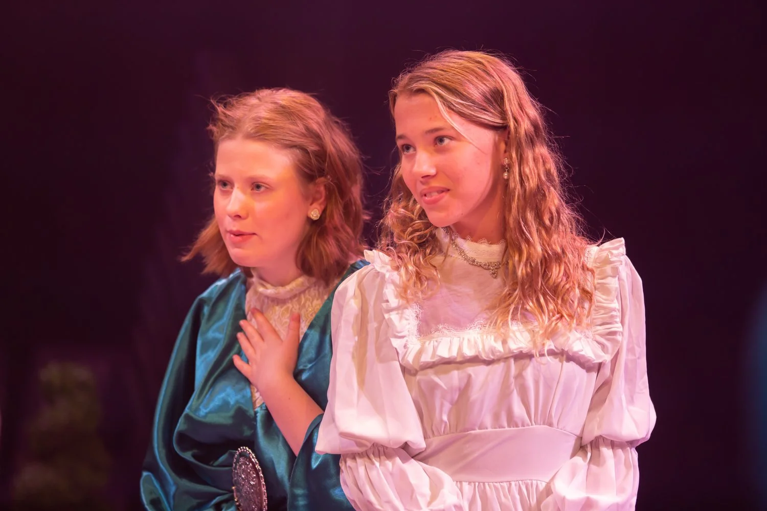 Two young girls dressed in vintage style costumes, standing closely together, performing on stage with a dark background.