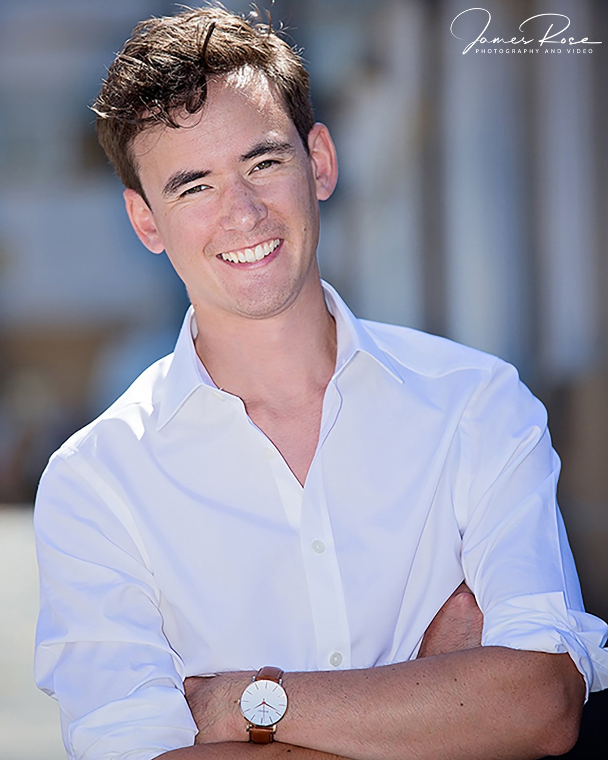 A young man with brown hair wearing a white shirt and a watch, smiling with arms crossed outdoors.