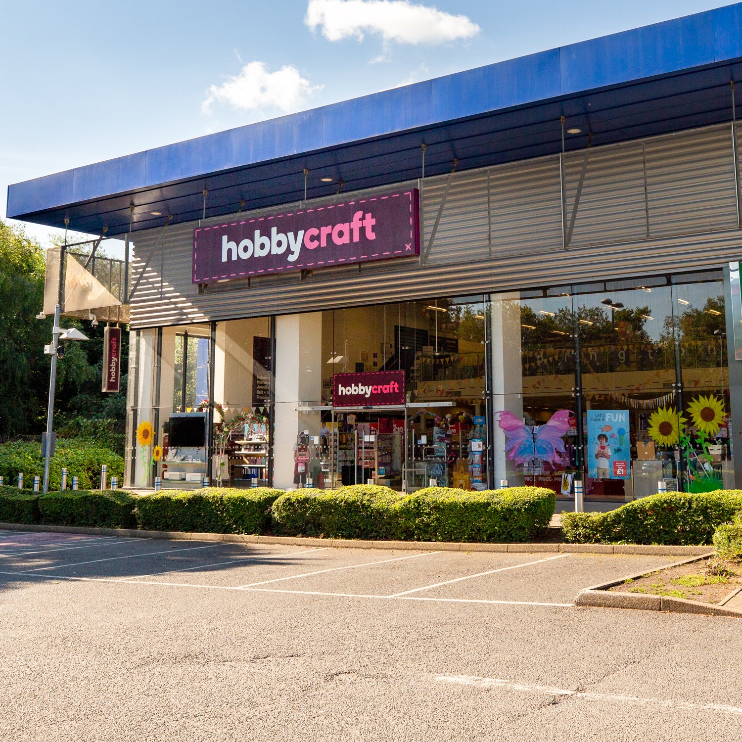 Exterior of a Hobbycraft store with large pink and white signage, glass windows showing inside displays, and a parking lot in front with trimmed bushes.