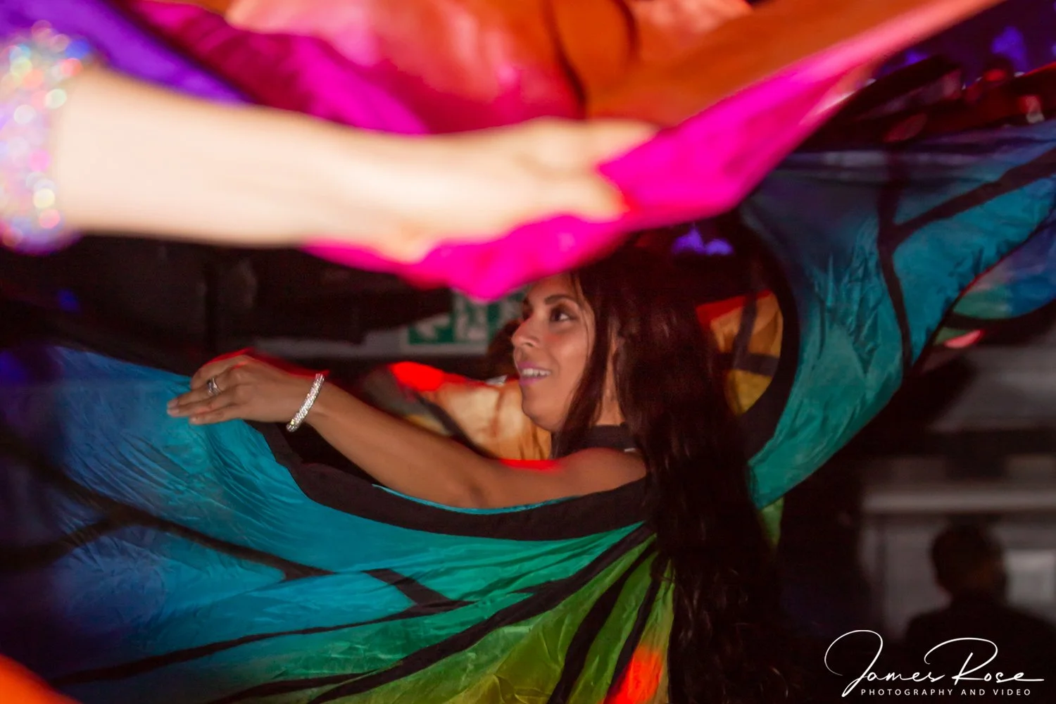 A woman in a colorful dress with vivid shades of pink, blue, green, and orange is dancing or spinning with an excited expression. She is wearing jewelry, including a bracelet and rings, and is holding part of her dress with her arm extended.