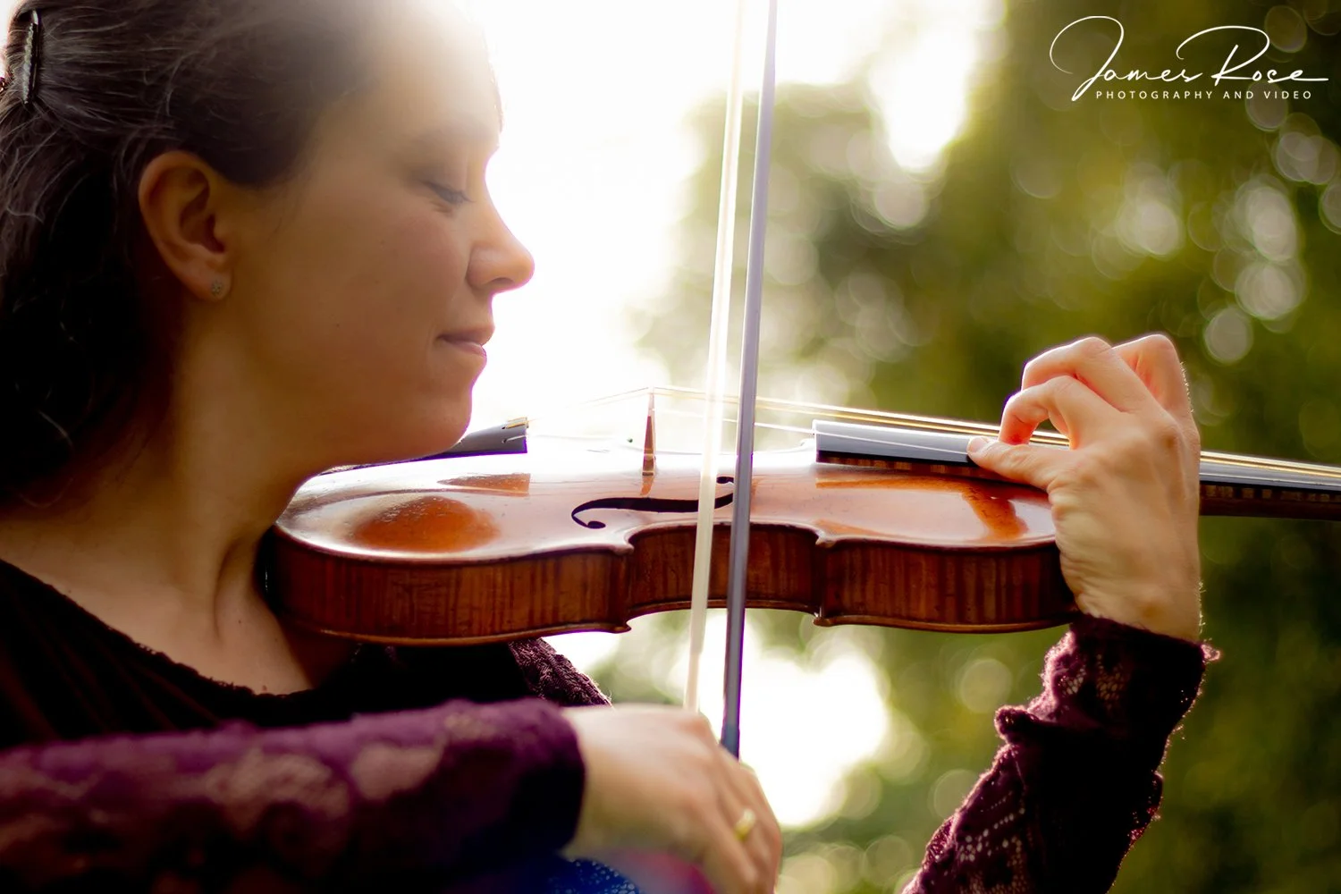 A woman playing the violin outdoors during daytime, with sunlight and blurred green background.