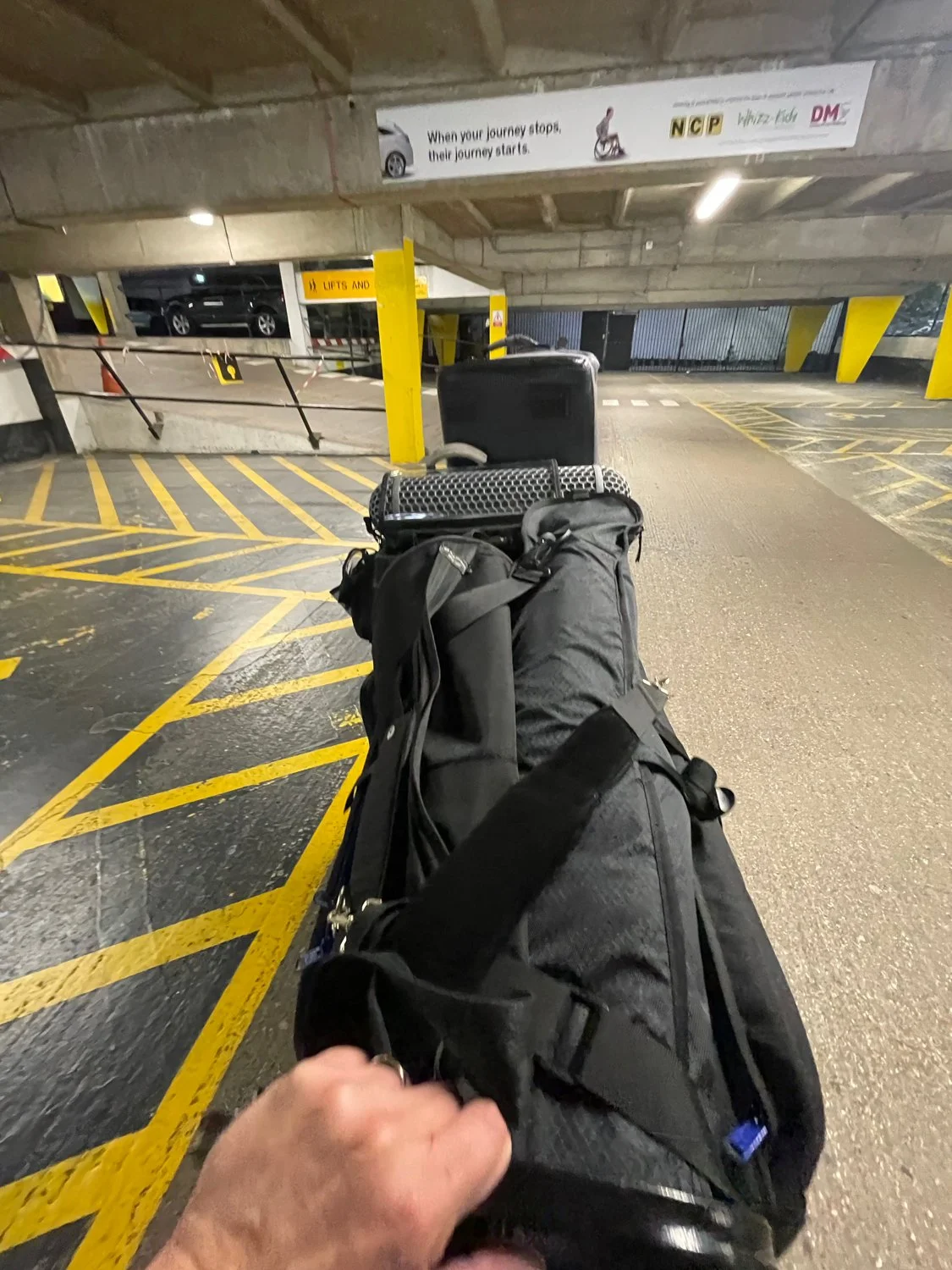 Person pulling a black rolling suitcase and carrying a black duffel bag in the parking garage.